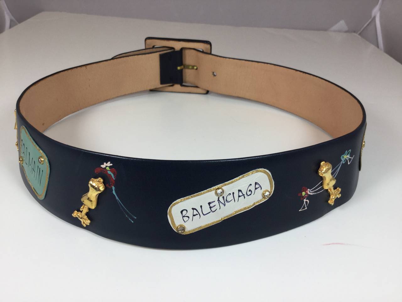 This delightful navy blue belt is decorated with tributes to all the major fashion houses of the day.  Each hand painted leather plaque is decorated with a designer name.  We have Dior, Fath, Balenciaga, Givenchy, and Balmain. The name plaques are