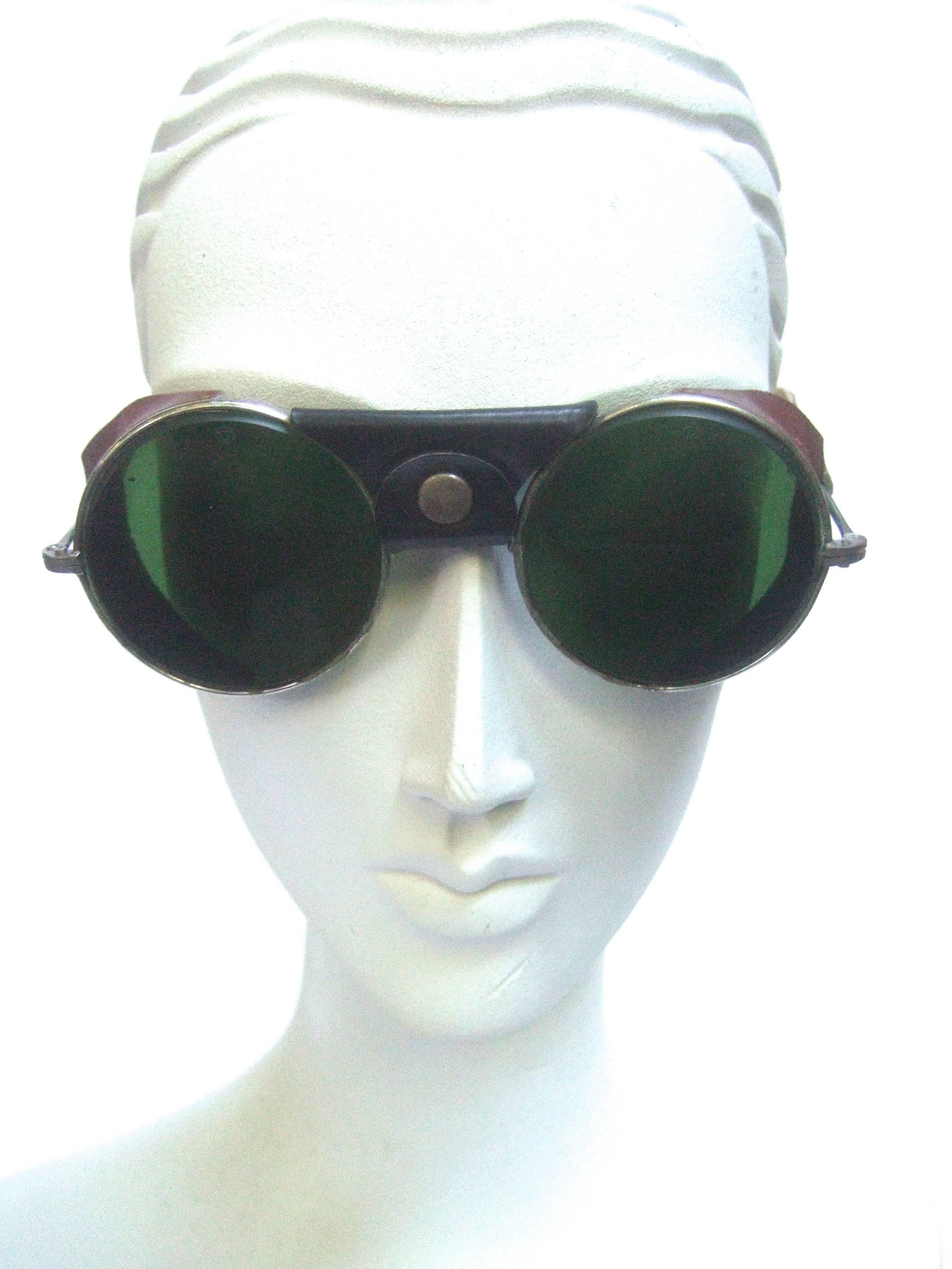 1940s Glass lens leather trim unisex aviator sunglasses 
The avant grade retro sunglasses are designed with 
circular dark green glass lenses 

The sides of the unique sunglasses are designed 
with brown leather perforated panels 
The center section