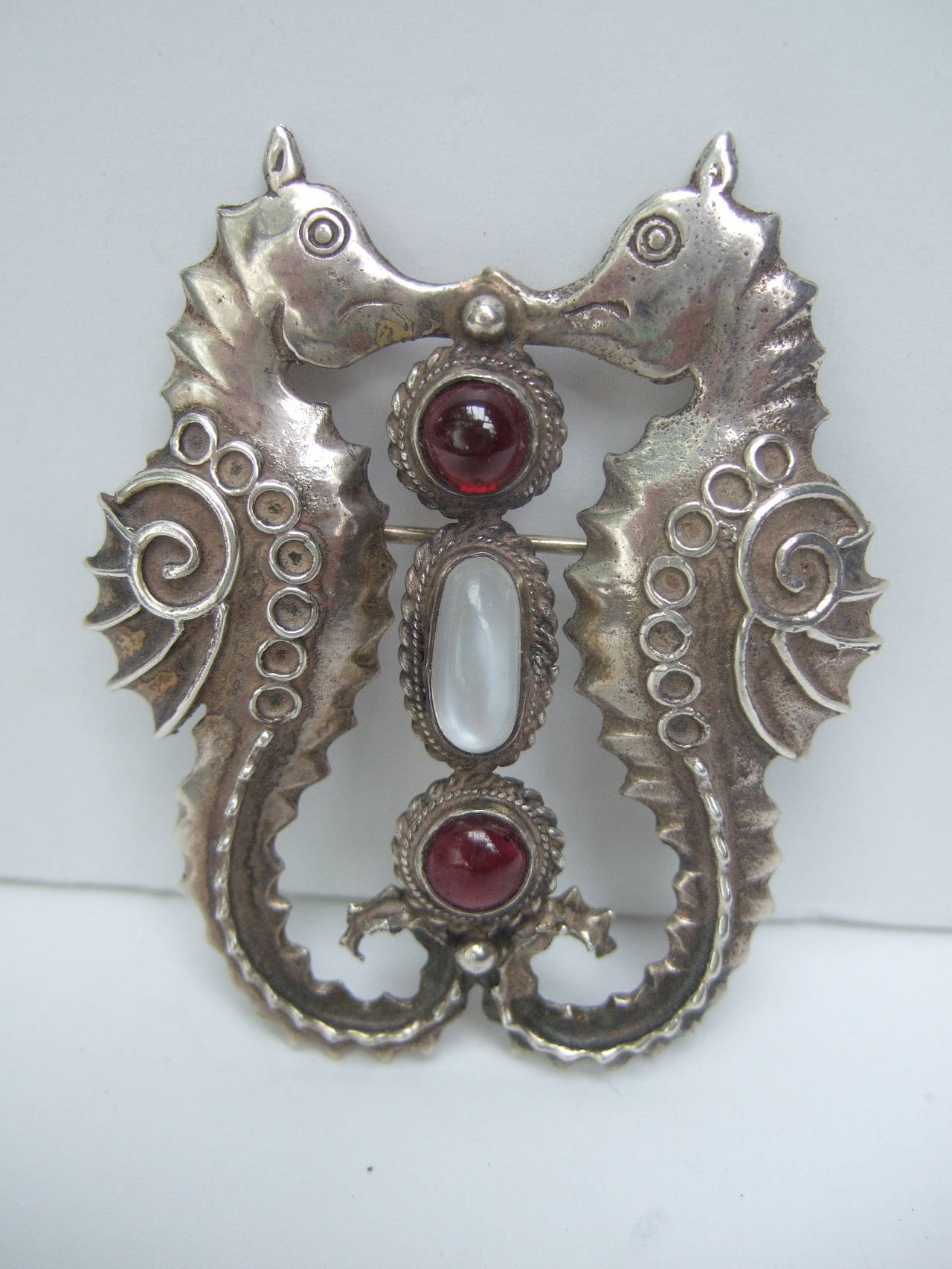 Sterling sea horse figural cabochon brooch c 1970
The handmade artisan brooch is designed with a
pair of stylized sea horses with scrolled circular 
stampings 

The center of the sterling brooch is embellished
with two garnet color round glass