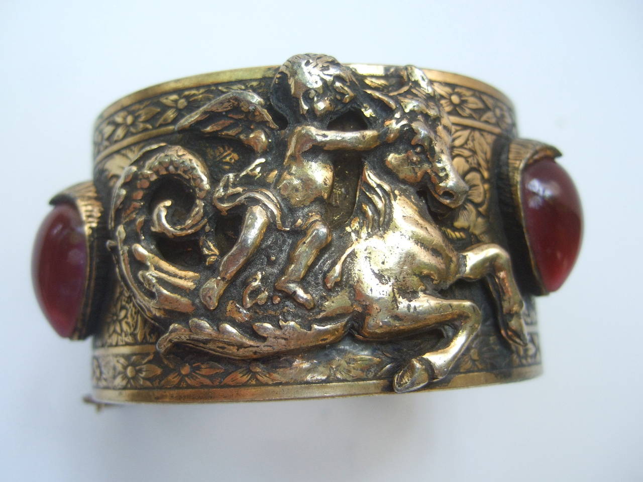 Ornate repousse gilt metal cherub cuff bracelet c 1970
The wide gilt metal cuff is adorned with a cherub 
figure riding on top of a mythical pegasus horse  

The wide gilt metal cuff is embellished with two deep
burgundy red glass streaked oval