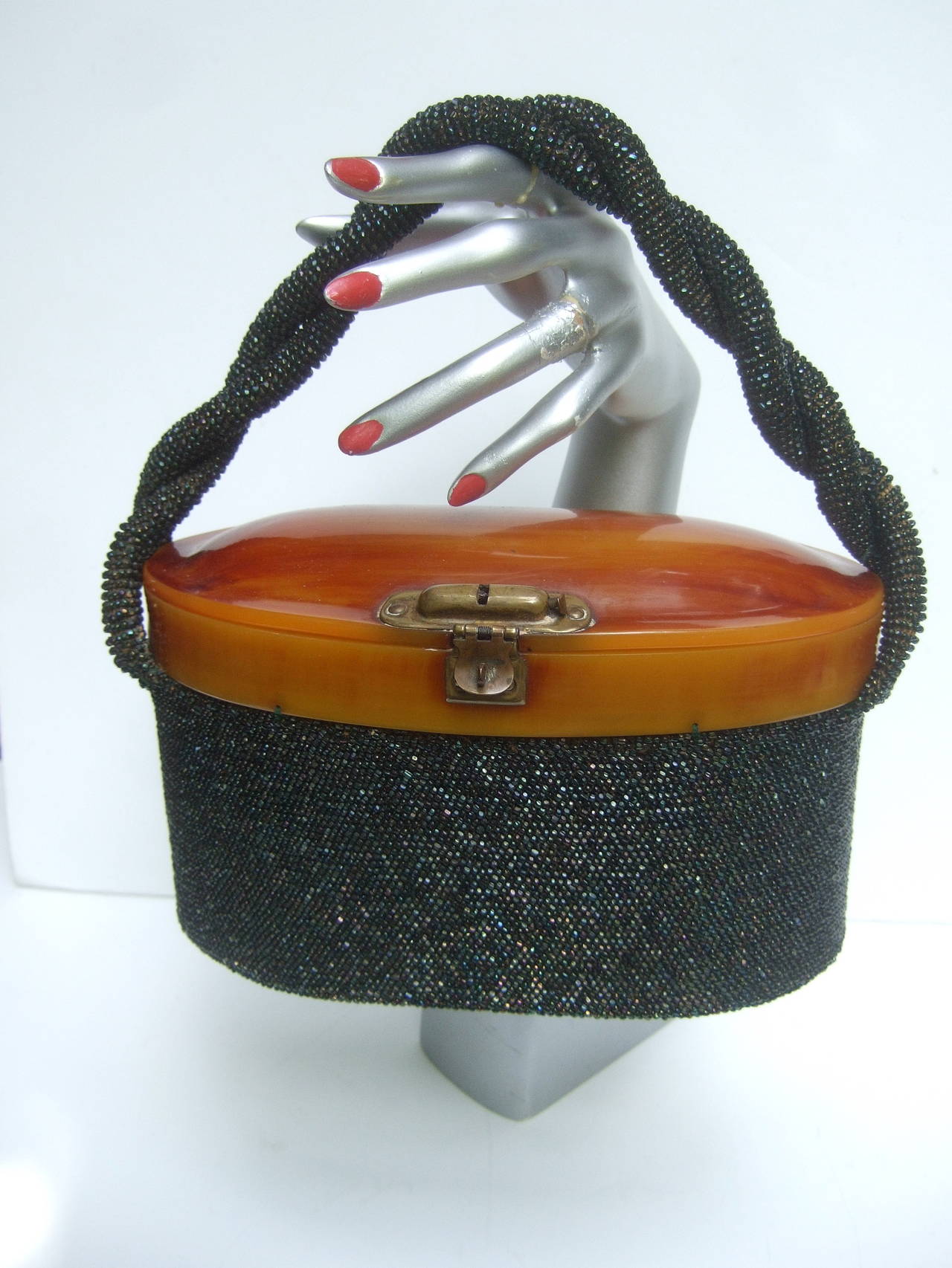 1940s Art Deco jet glass beaded handbag 
The oval shaped carnival beaded is adorned
with an amber lucite lid cover. The elegant 
retro handbag is designed with a brass metal
latch clasp mechanism 

The black glass micro beading circles the