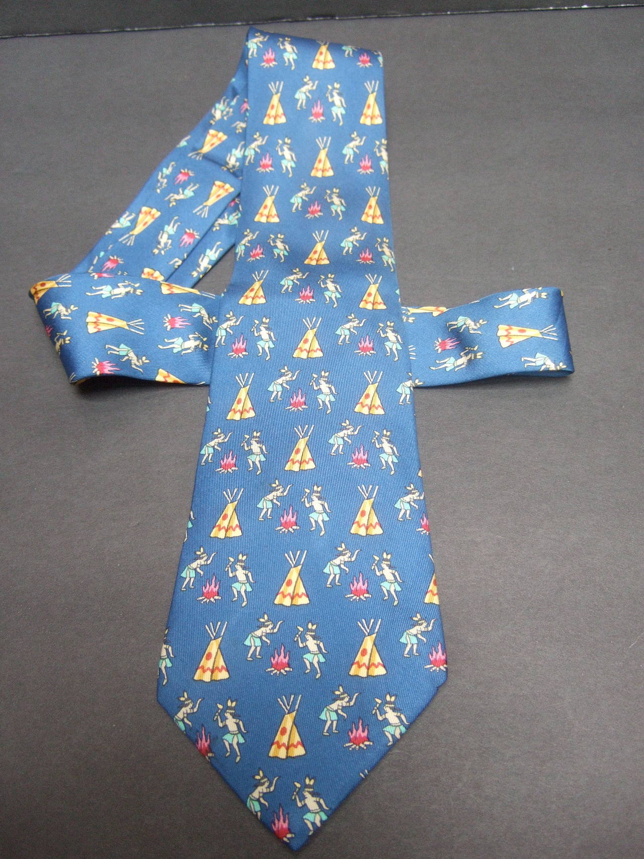 Hermes Paris Whimsical American Indian silk print necktie c 1990s
The unique designer silk necktie is illustrated with pairs 
of warriors dancing around a burning fire. The cobalt blue
background is designed with teepee huts

Labeled Hermes