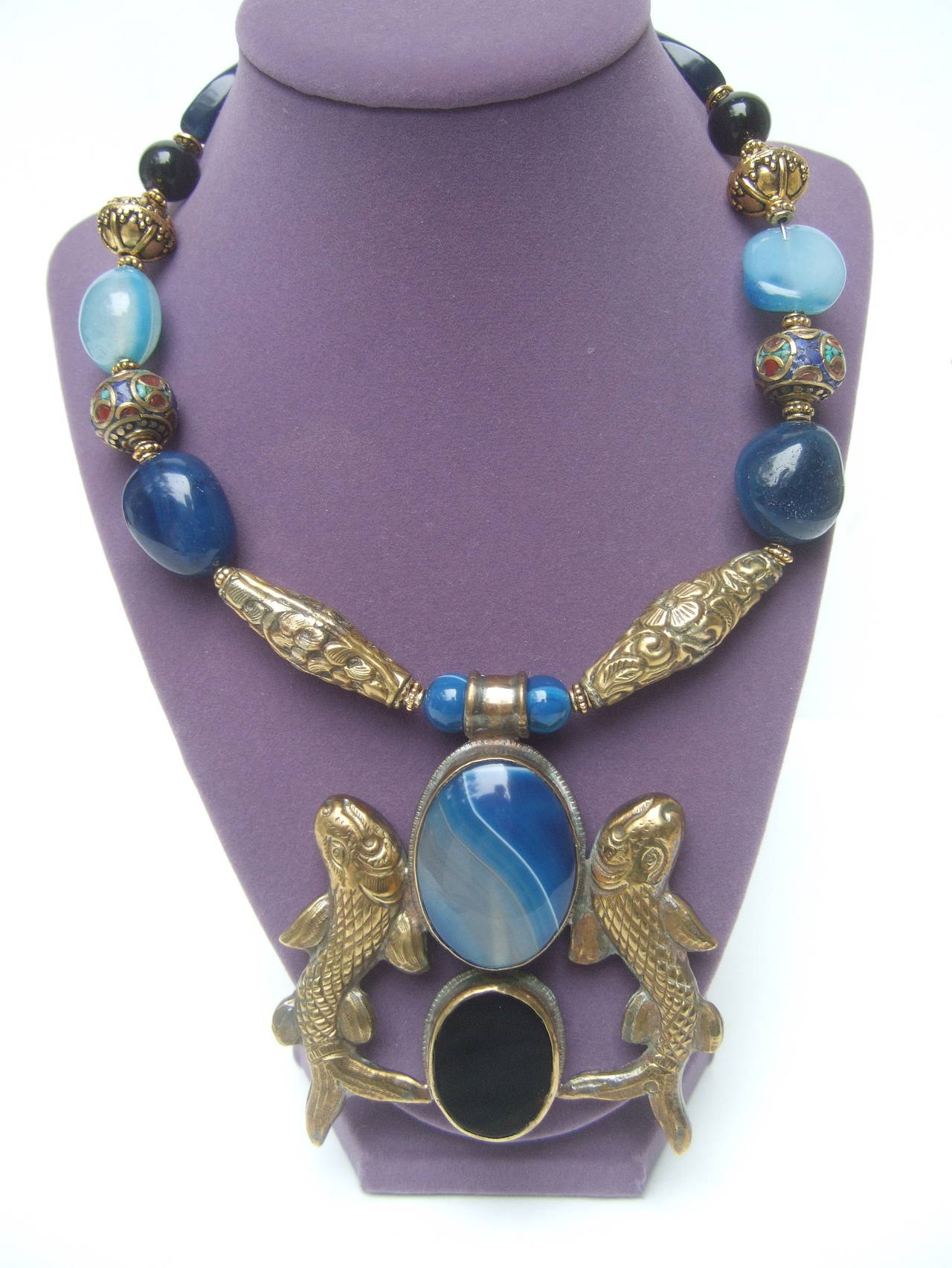 Exotic artisan Nepalese glass stone choker necklace c 1980s
The unique handmade necklace is designed with a 
pair of large brass metal fish. The fish pendant is 
adorned with a large blue agate oval shaped cabochon

Under the large blue agate