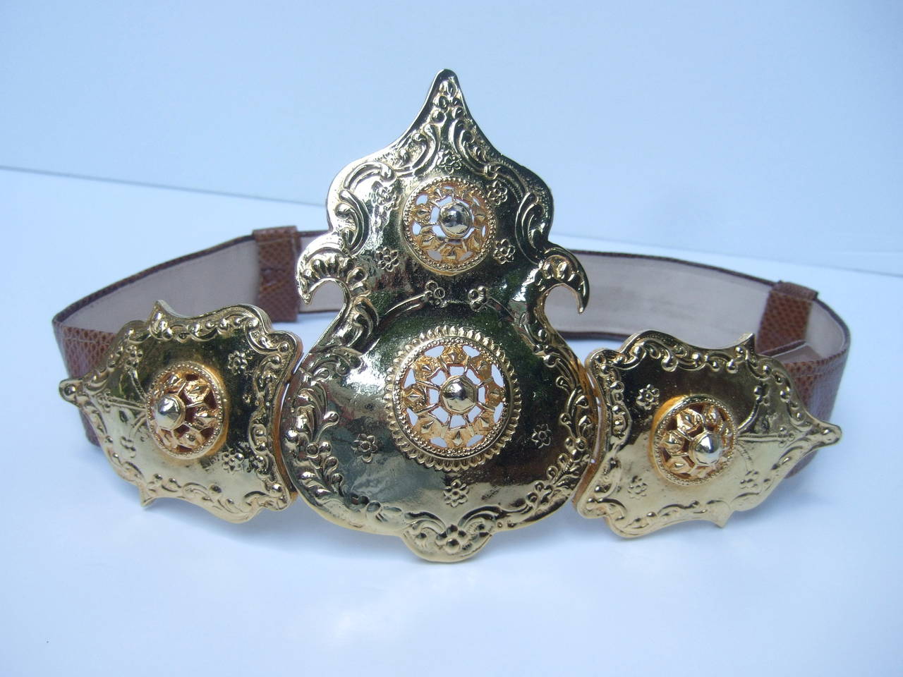 ALEXIS KIRK Massive ornate gilt metal buckle brown leather belt c 1980s
The designer belt is adorned with a huge gilt metal gladiator style buckle that is embellished with scrolled repousse designs 

The brown leather adjustable belt is designed