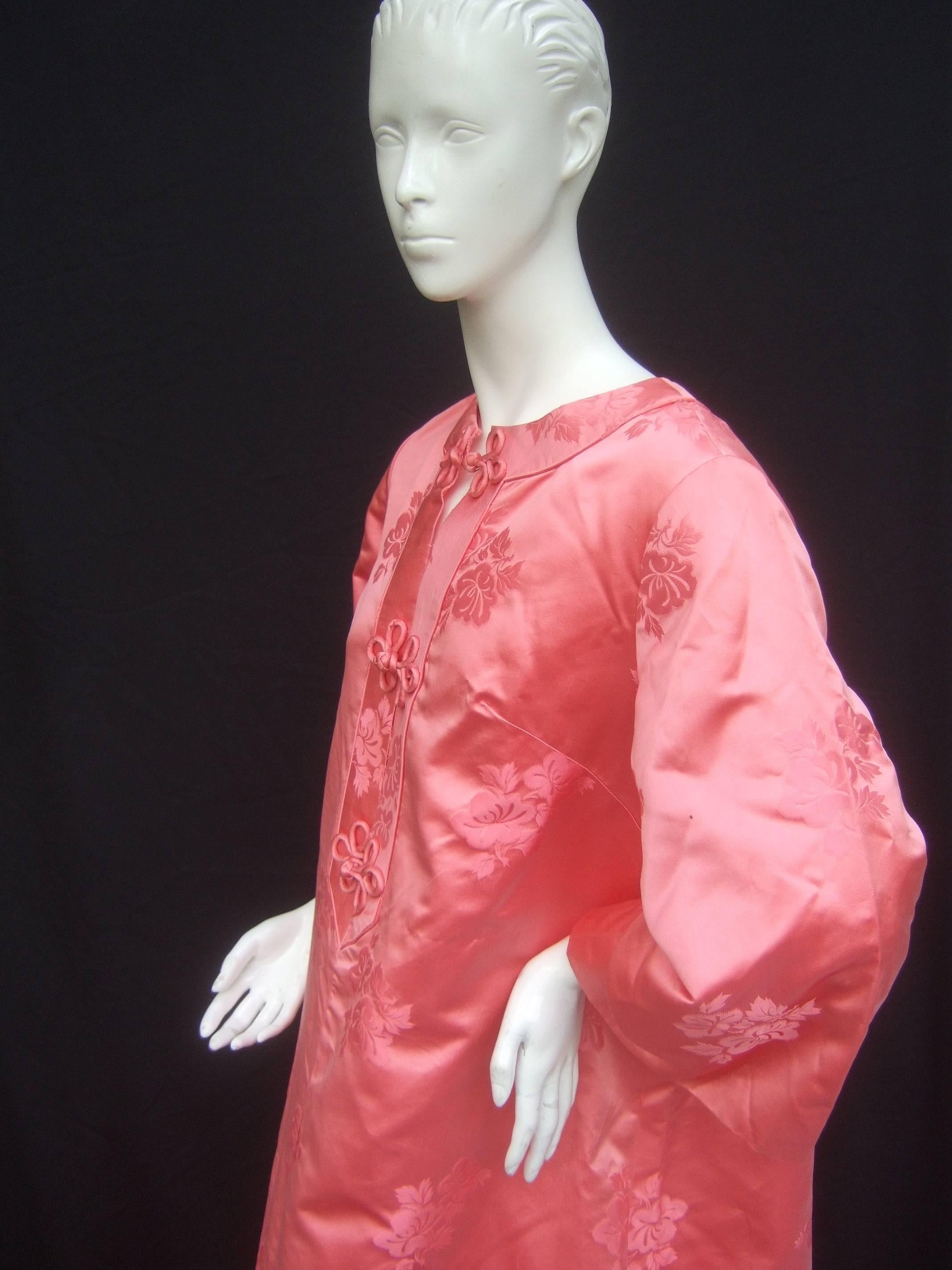 Coral pink satin damask caftan gown c 1970s
The Asian inspired caftan is designed with 
luminous pink satin with recurring subtle 
floral designs

The neckline is adorned with frog closures 
that run down the bodice. The luxurious 
caftan is lined