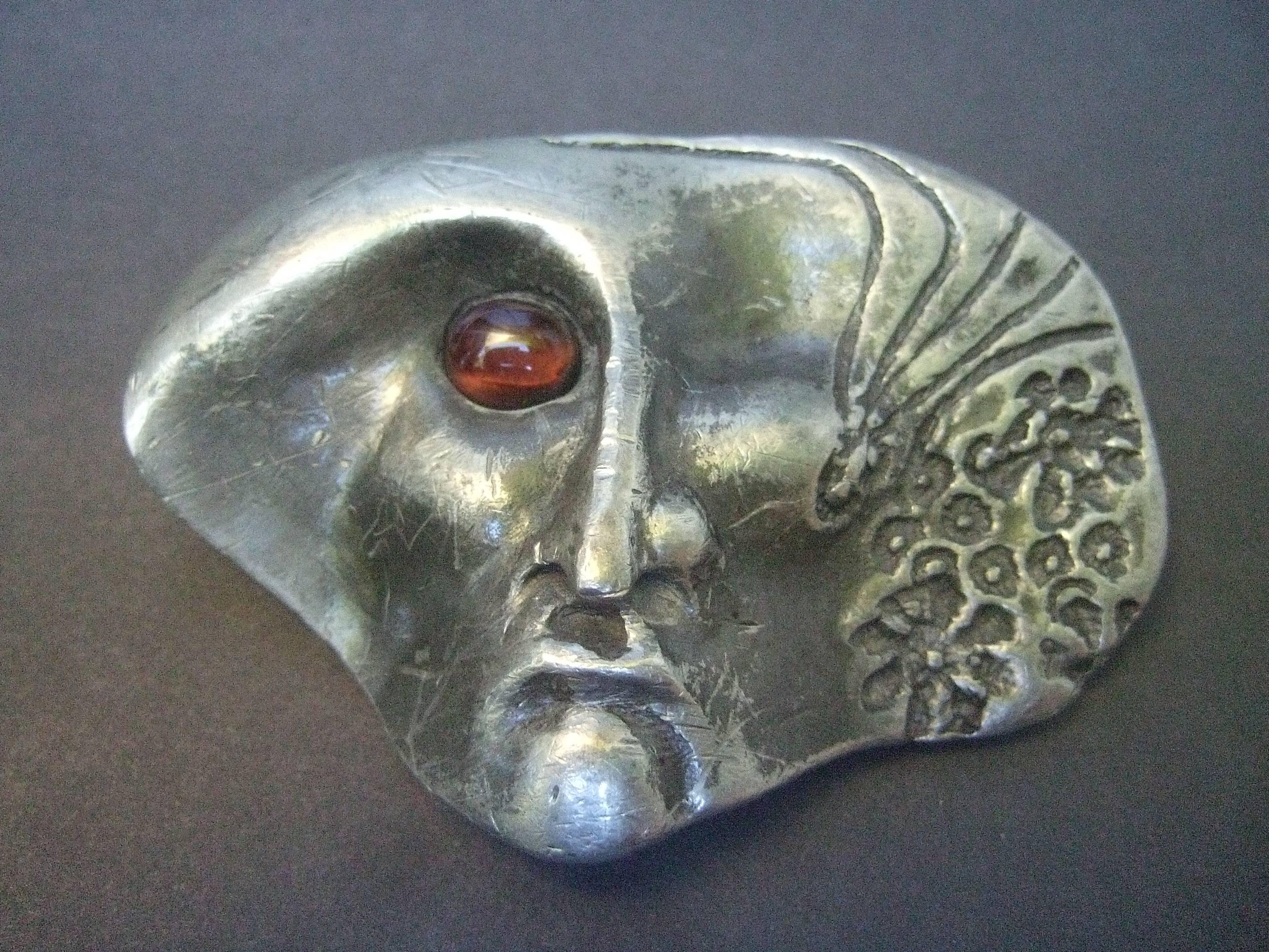 Avant-garde silver metal artisan belt buckle by Lynne Dorsch 
The unique belt buckle is designed with a surrealistic 
face. The face is adorned with a smooth glass cabochon
that distinguishes one eye 

One side of the figures face is