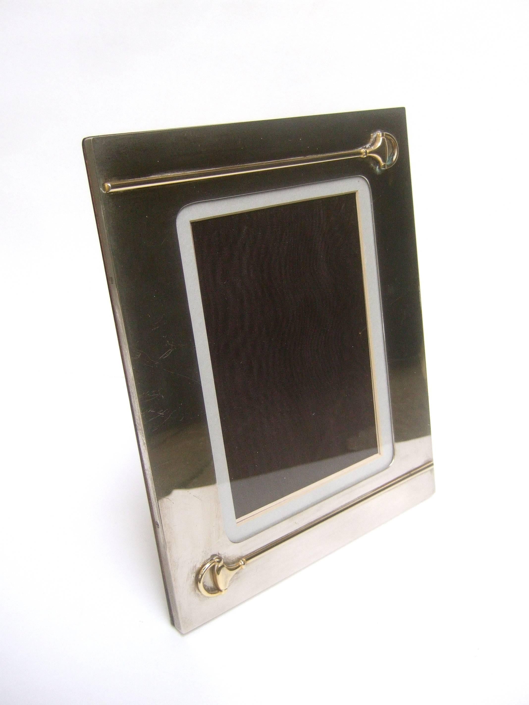 Gucci Italy Sleek chrome metal picture frame c 1970s 
The elegant chrome photo frame is embellished 
with a pair of Gucci's signature gilt metal bridals

The interior backing is taffeta. The back exterior
side is lacquered wood. The base is