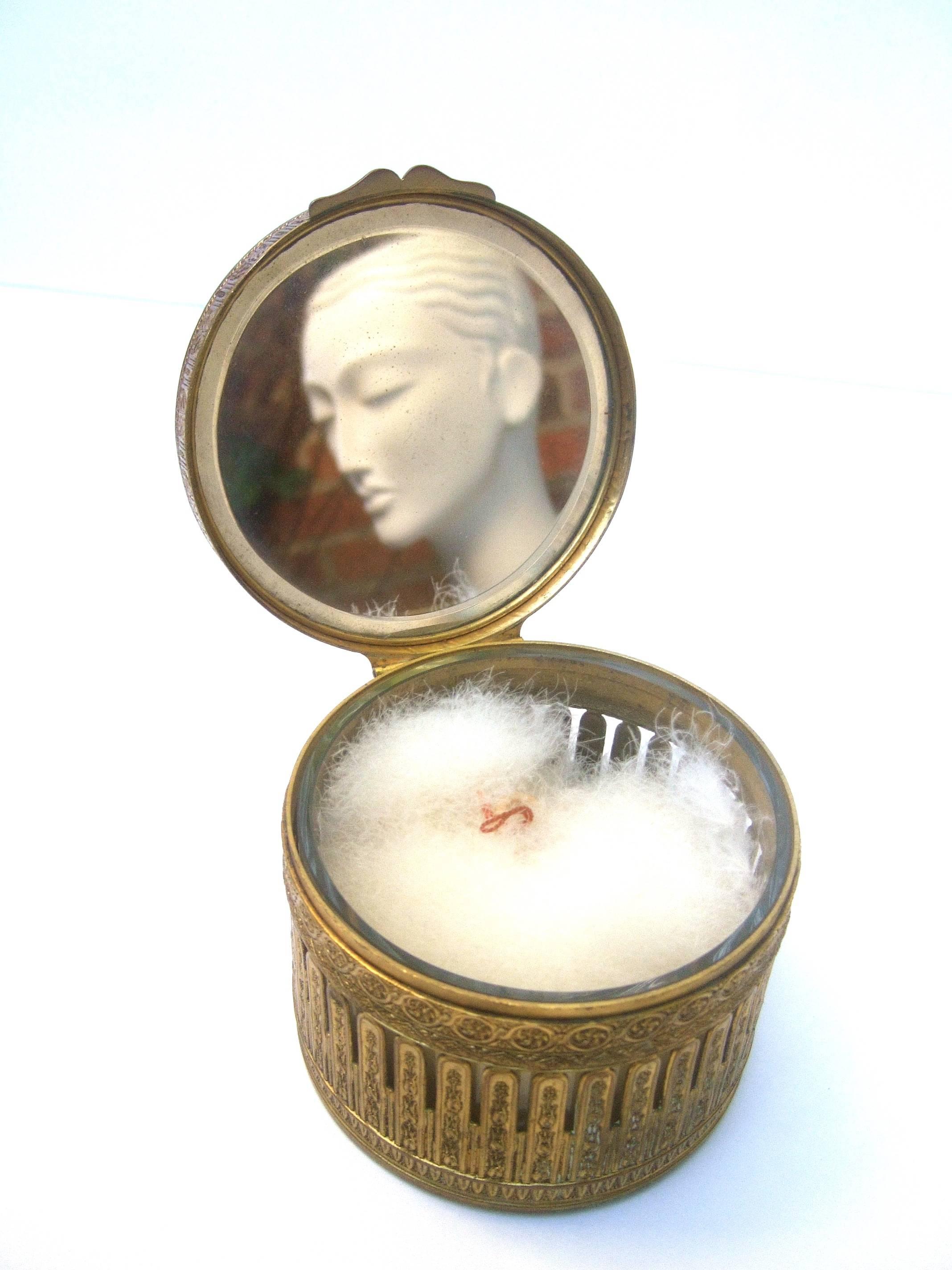 French gilded metal powder puff vanity trinket box
The ornate round brass metal box is decorated
with intricate floral motifs throughout

The center of the lid cover depicts a portrait 
style image of an elegant woman under a clear
lucite cover. The