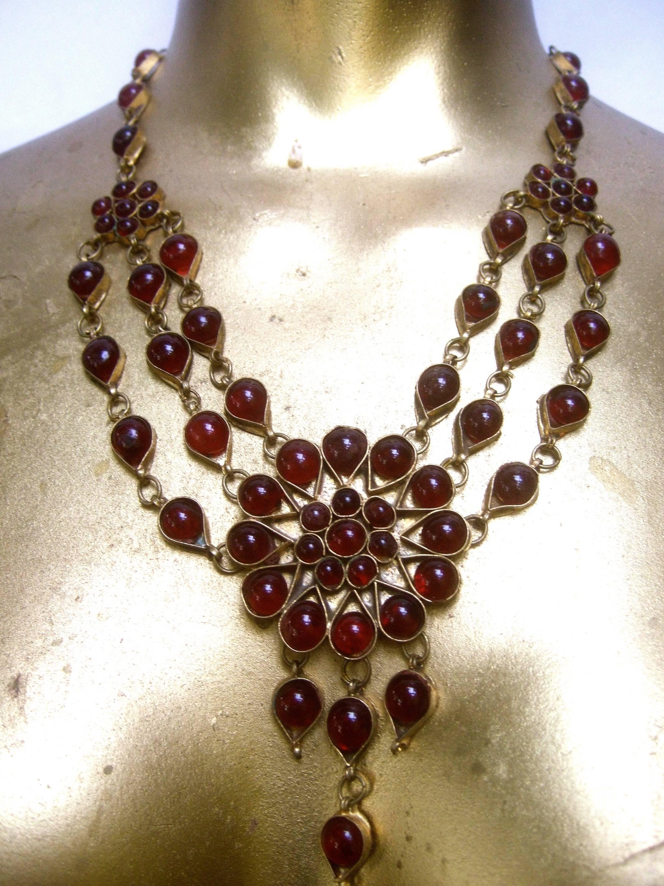 Exotic cranberry glass cabochon choker necklace 1980s
The ethnic artisan necklace is embellished with clusters
of deep red glass stones

The center circular medallion is designed with three
rows of dangling glass settings. The sides transition
into