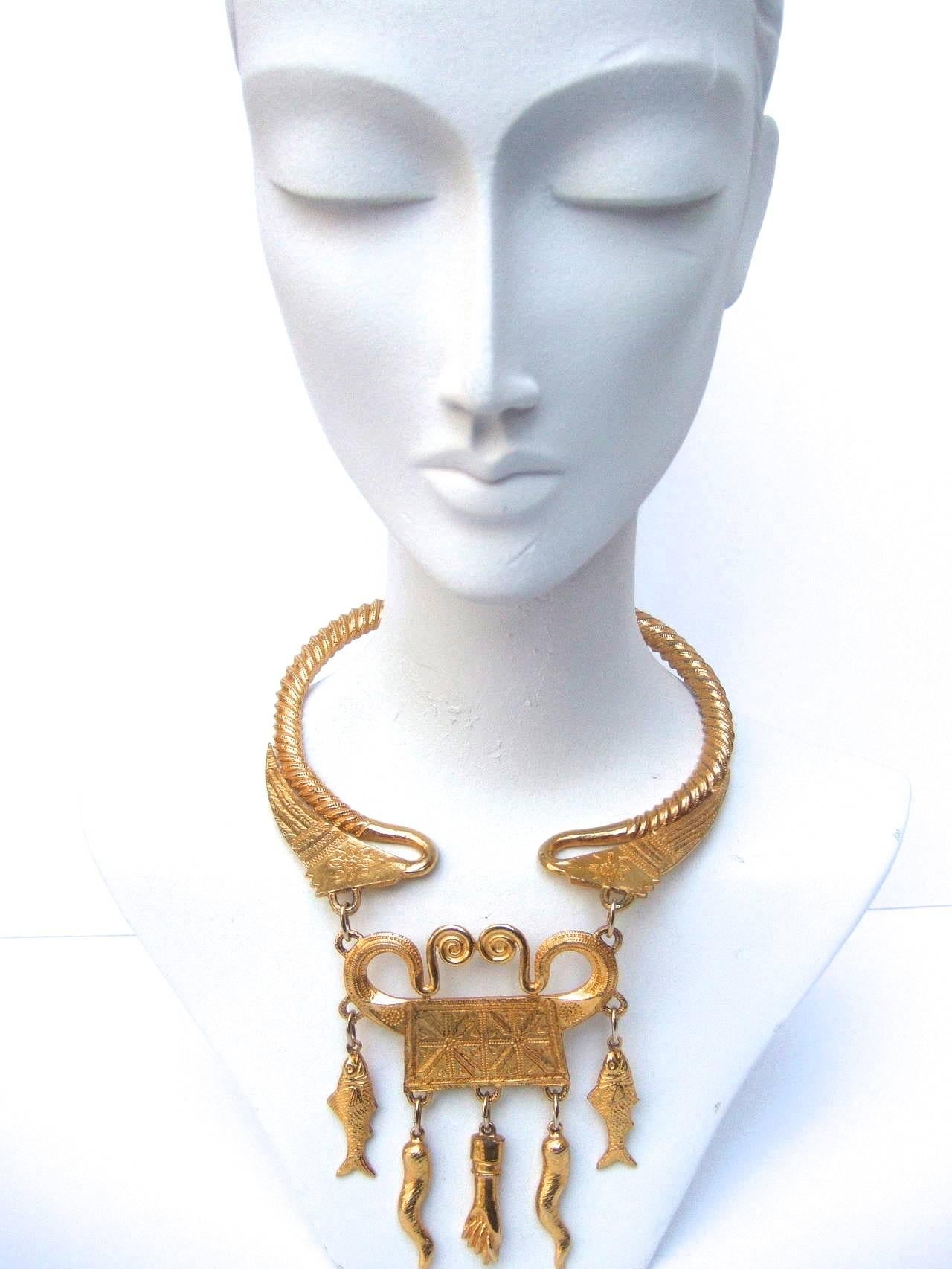 This exotic large scale necklace by Alexis Kirk is designed with dangling horns, fish, and hand embellishments suspended from a central plaque.

The gilded choker is made of chunky, very solid feeling, metal with grooved etchings.  The front edges