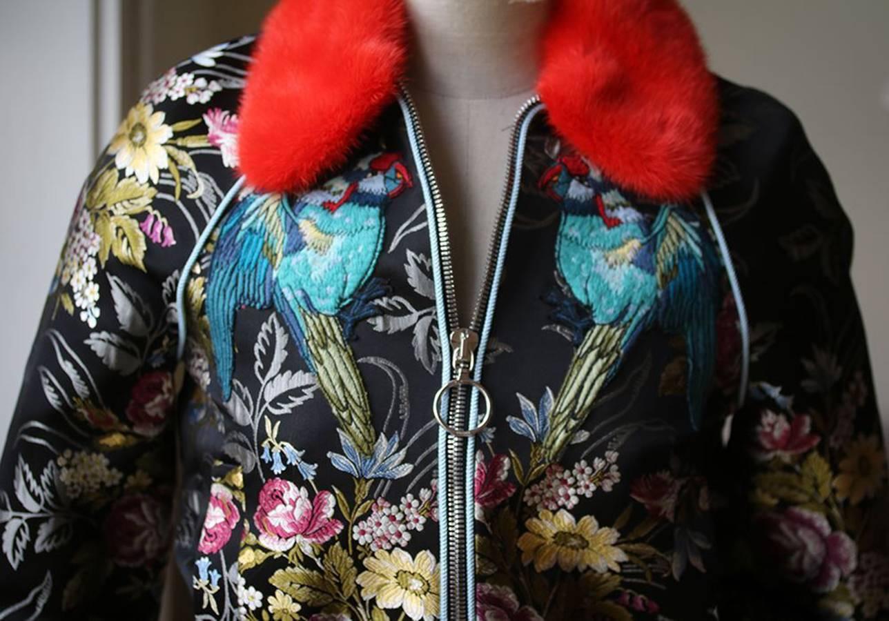 A floral jacquard bomber jacket features with animal embroidery at the front and back, including the parrot and the butterfly. Black floral silk jacquard. Orange dyed mink fur (Finland) collar. Embroidered parrot and butterfly appliqué. Knit Lurex®