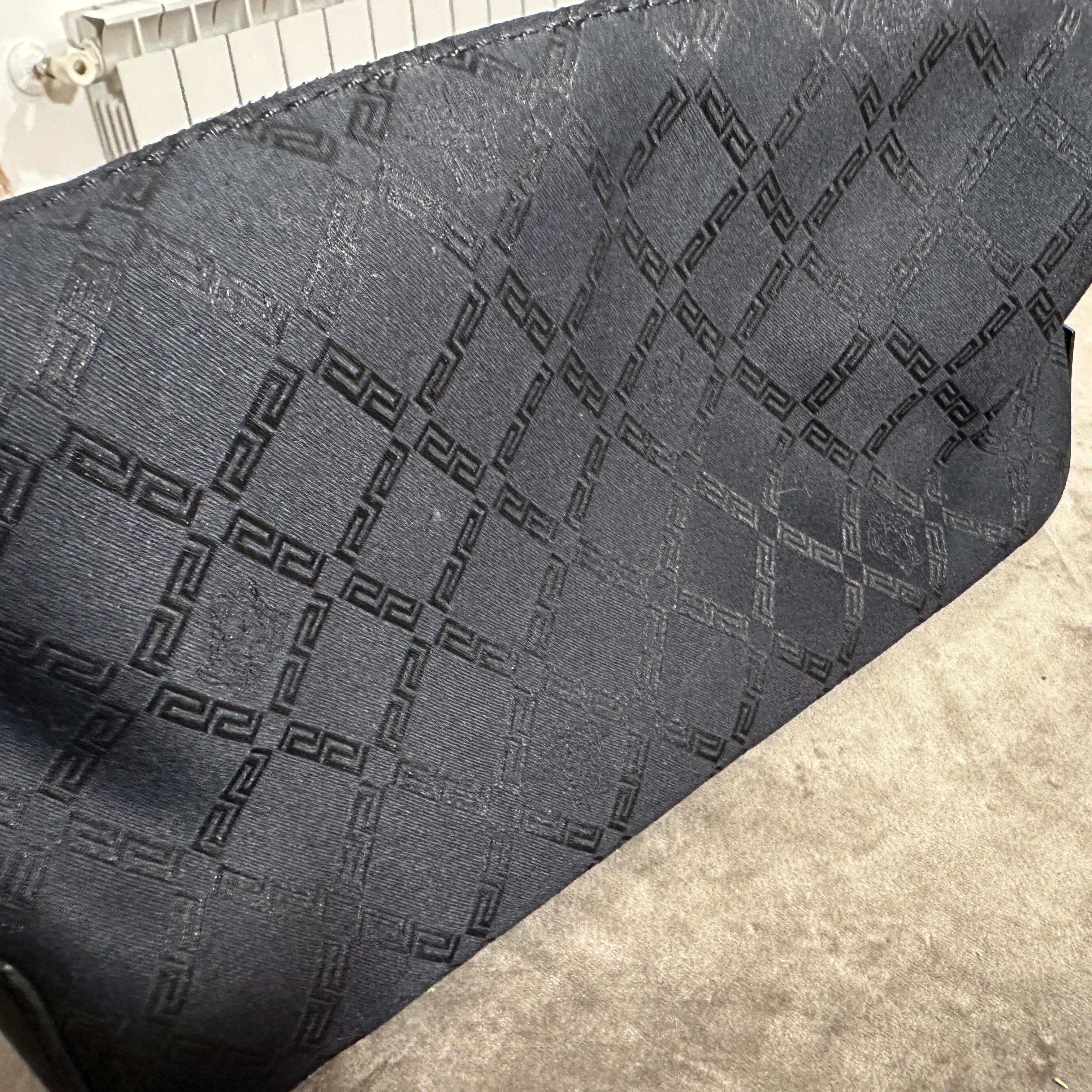 Entdecken Sie diese schwarze Handtasche von Gianni Versace aus den 1990er Jahren, ein raffiniertes Stück, das die kühne und zugleich elegante Ästhetik dieser Zeit verkörpert. Es ist aus strapazierfähigem schwarzem Stoff gefertigt und weist ein