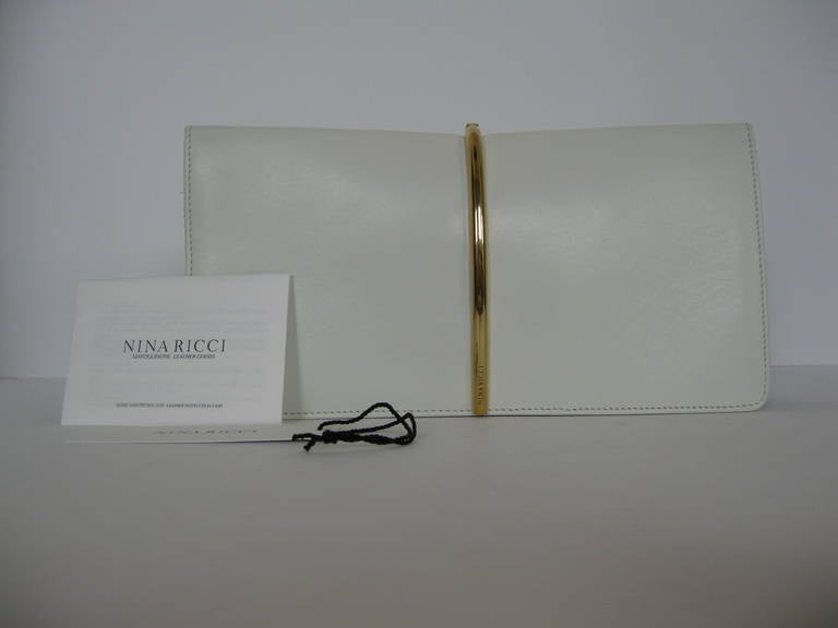 NINA RICCI white calfskin Arc Clutch.
Arc Clutch styled over the top line with a gold tone metal arc. 
Front flap, logo engraved goldtone metal hinged arc. Black suede gusseted sides.
Lined in natural leather with a slip pocket.
Magnetic