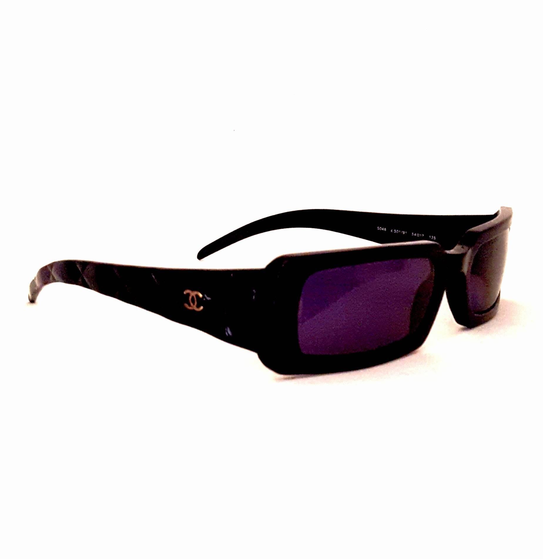 Les lunettes de soleil rectangulaires de Chanel sont dotées du matelassage classique de Chanel sur la longueur de la branche ainsi que de petits logos CC argentés de chaque côté.  Cette paire est en plastique noir avec des verres polarisés gris