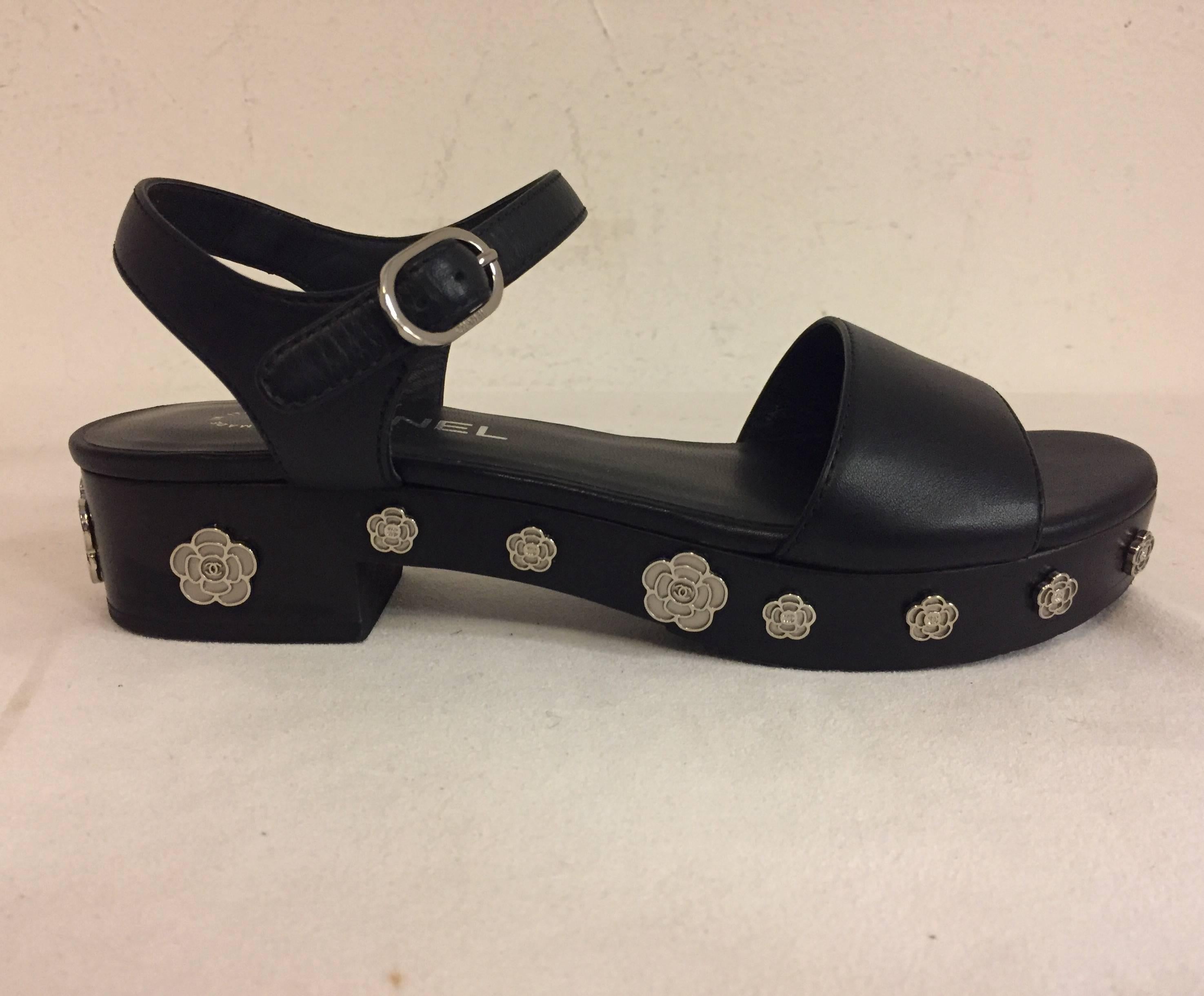 These creative Chanel sandals with platforms are simply crafted of calfskin leather in black.  The feature black wooden heel and platform lined with silver tone Camellia flowers.  The sandal has a wide single strap across the toes and an adjustable