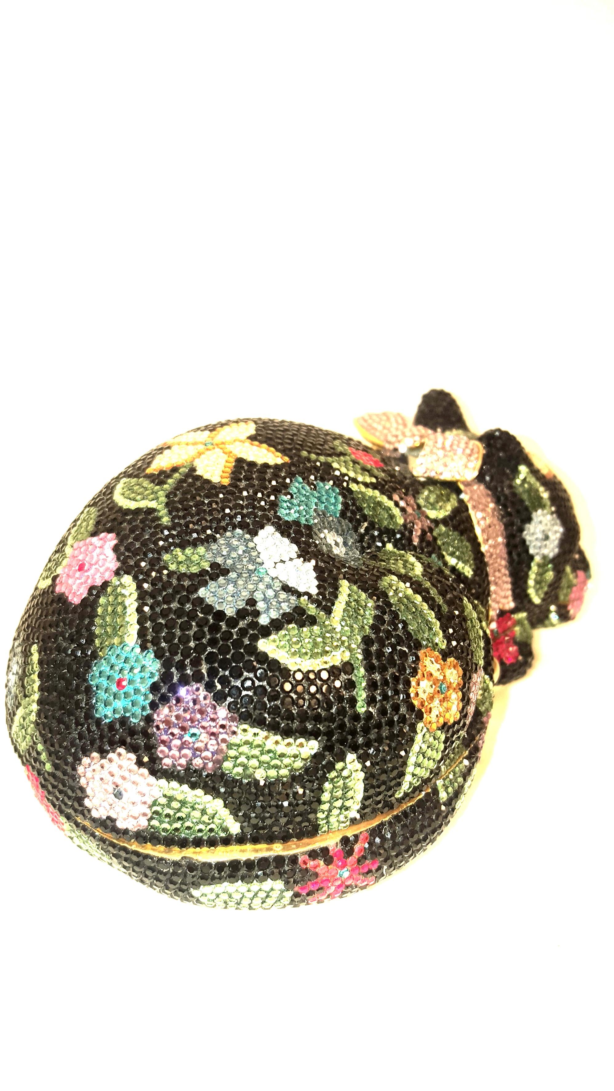 Judith Leiber minaudière features the form of a bejeweled sleeping cat with a bow around its neck. The hard clutch is covered all over with mini Swarovski crystals with black background and multi flowers floating around in shades of yellow, red,