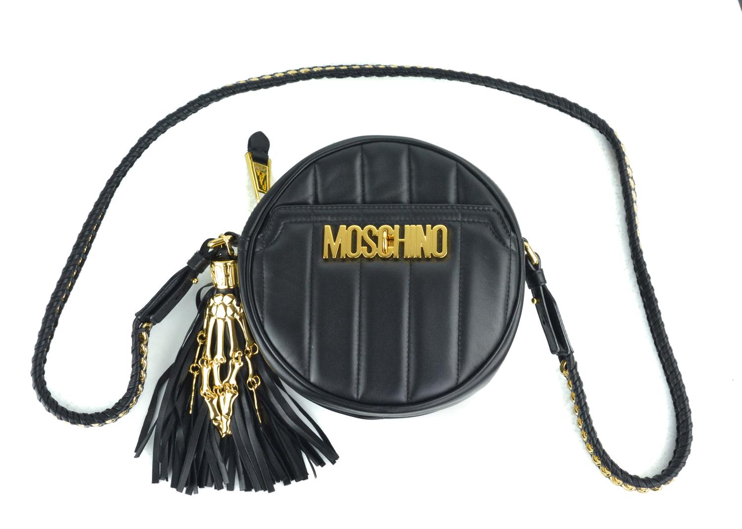Moschino's black leather quilted round shoulder bag. This bag features a gold tone hardware moschino logo with vertical quilted detailing. The bag also features a fringe onthe side with a gold tone skeletal hand to accompany it. Perfect everyday bag