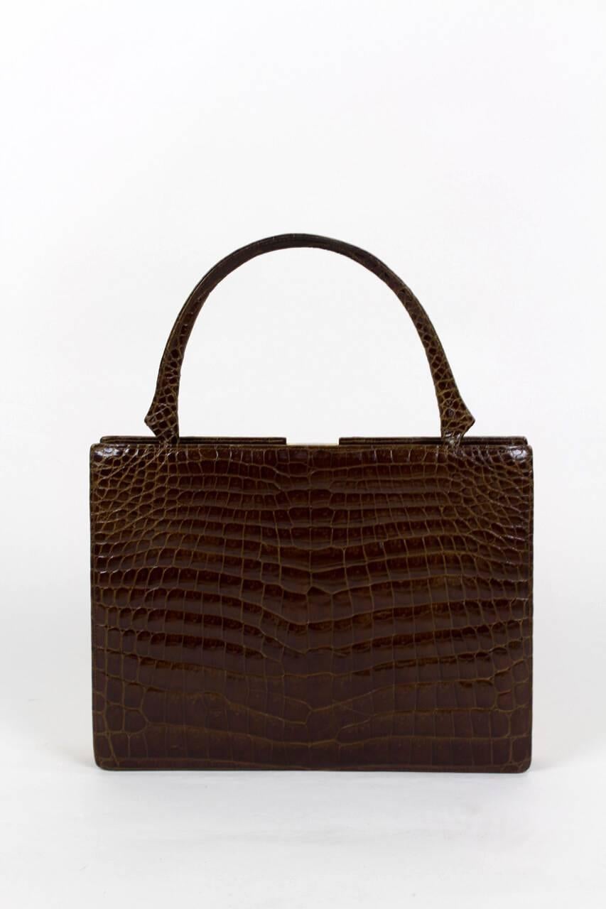 This crocodile print leather top handle bag shows a beautiful colour gradient from beige (matt central panel) to brown (glossy sides) on the front whereas the glossy back is monochrome in brown. It has a rectangular shape, a gold-tone frame with top