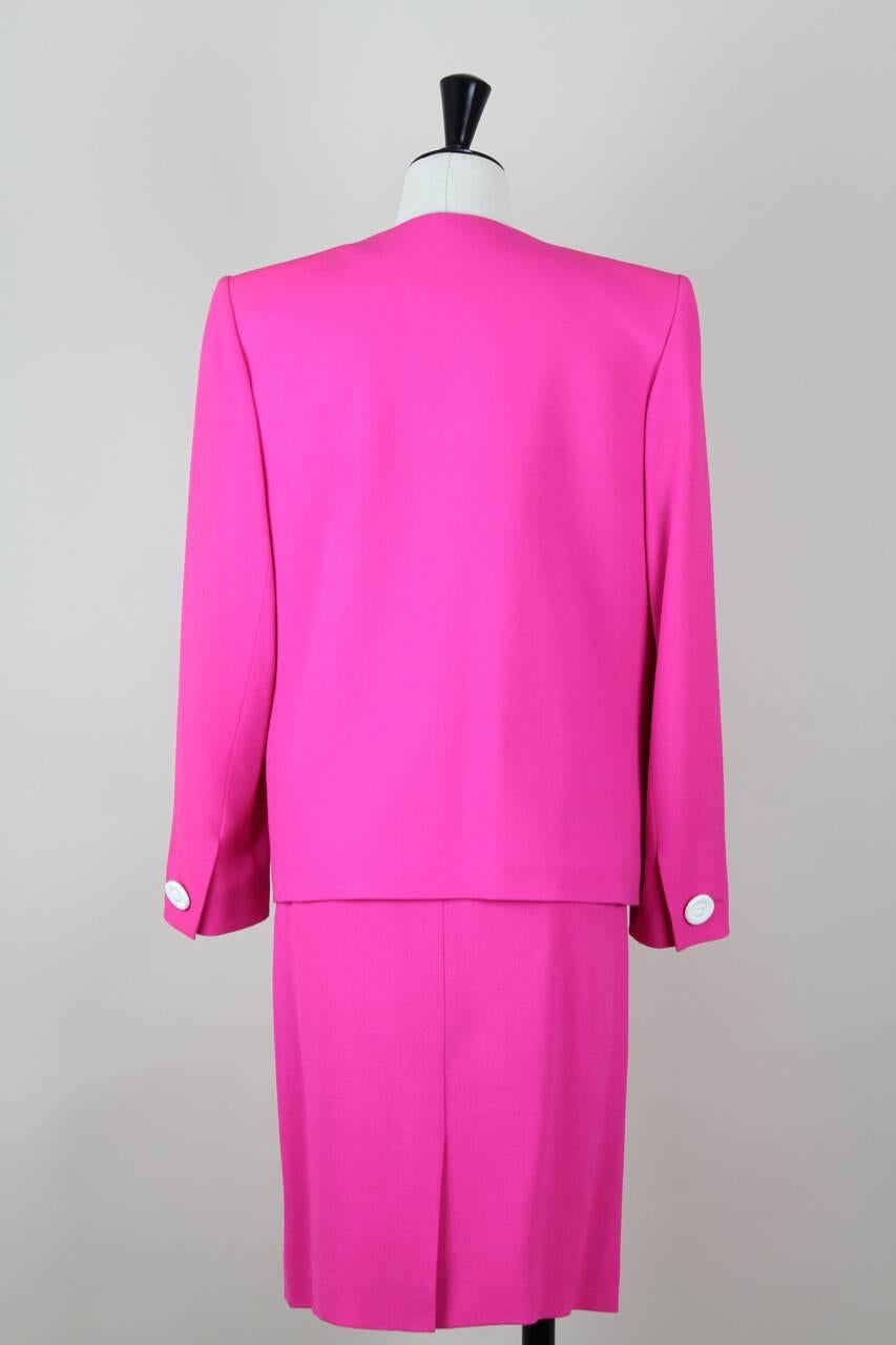 This jacket and skirt suit is made from a lightweight hot pink wool blend crepe. The collarless jacket has a box cut with shoulder pads, long sleeves with buttoned cuffs and two lower front lined patch pockets with an eye-catching white