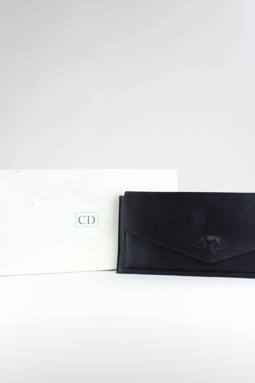 This envelope style clutch bag is made from black duchesse silk. It features a fold-over front flap decorated with a small bow and fastens with a snap. The bag is fully lined with black fabric, has two compartments with two side pockets (one in