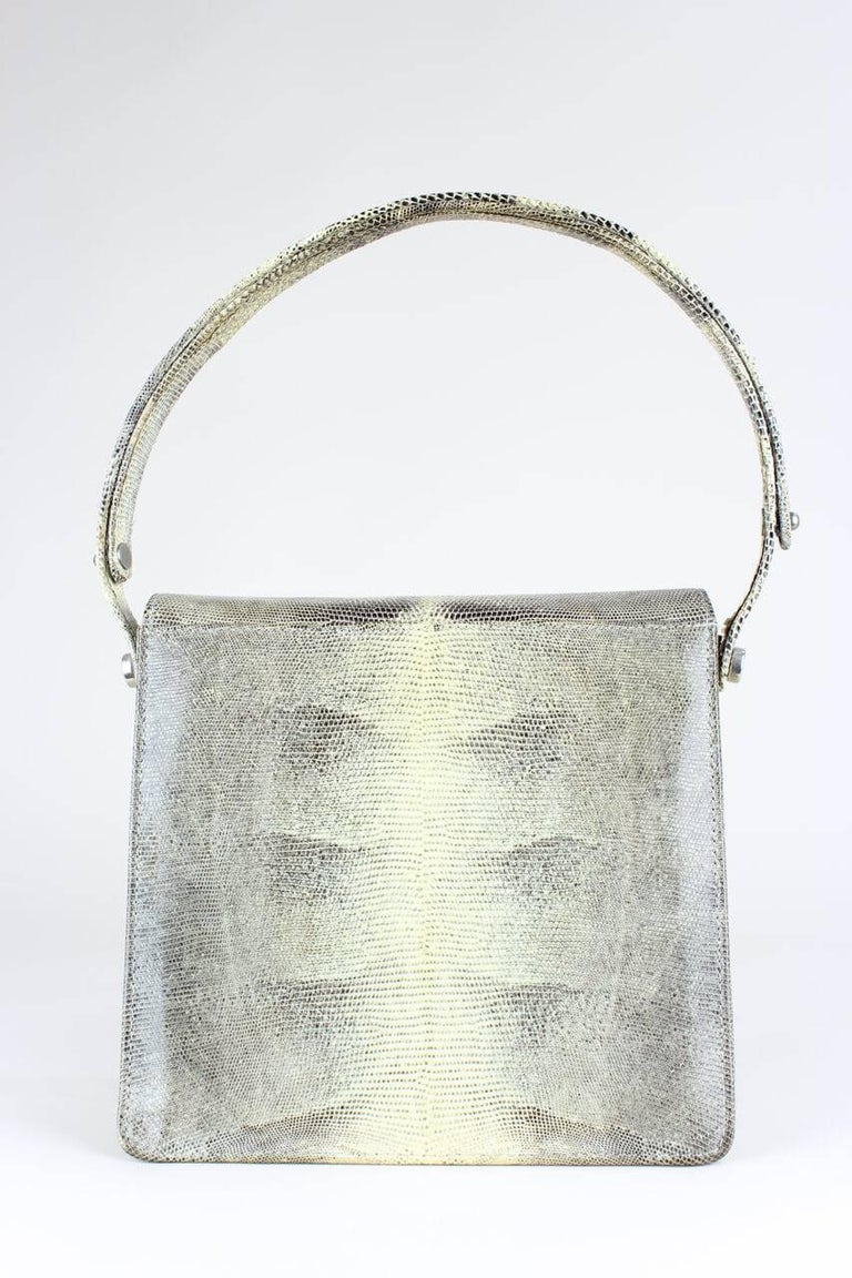 This square shoulder bag in greys and cream is made from lizard pattern leather. It features a fold-over front flap with a silver-toned push-lock fastening and a matching shoulder strap. The bag is fully lined in ivory-coloured leather and has two