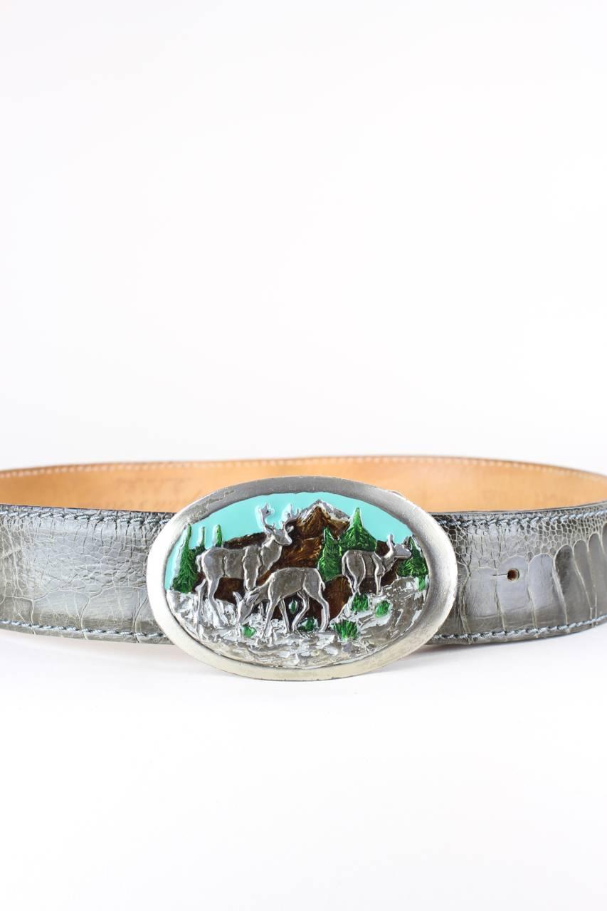 This lovely heavy and solid oval-shaped belt buckle is made from silver-toned metal. The buckle features an enamel design of deer in the mountains with turquoise sky, brown mountains and green firs and grass. The back has fine engraving details and