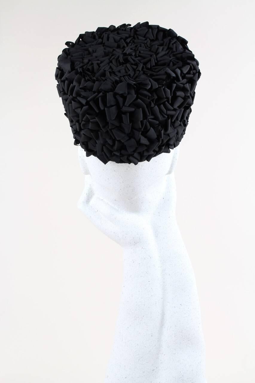 Dies ist ein ungewöhnlicher und raffinierter handgefertigter schwarzer Pillenbox-Hut aus den 1960er Jahren. Die Basis besteht aus schwarzem, versteiftem Tüll, der vollständig mit schwarzen, von Hand aufgenähten Taftschleifenbändern bedeckt ist. Die
