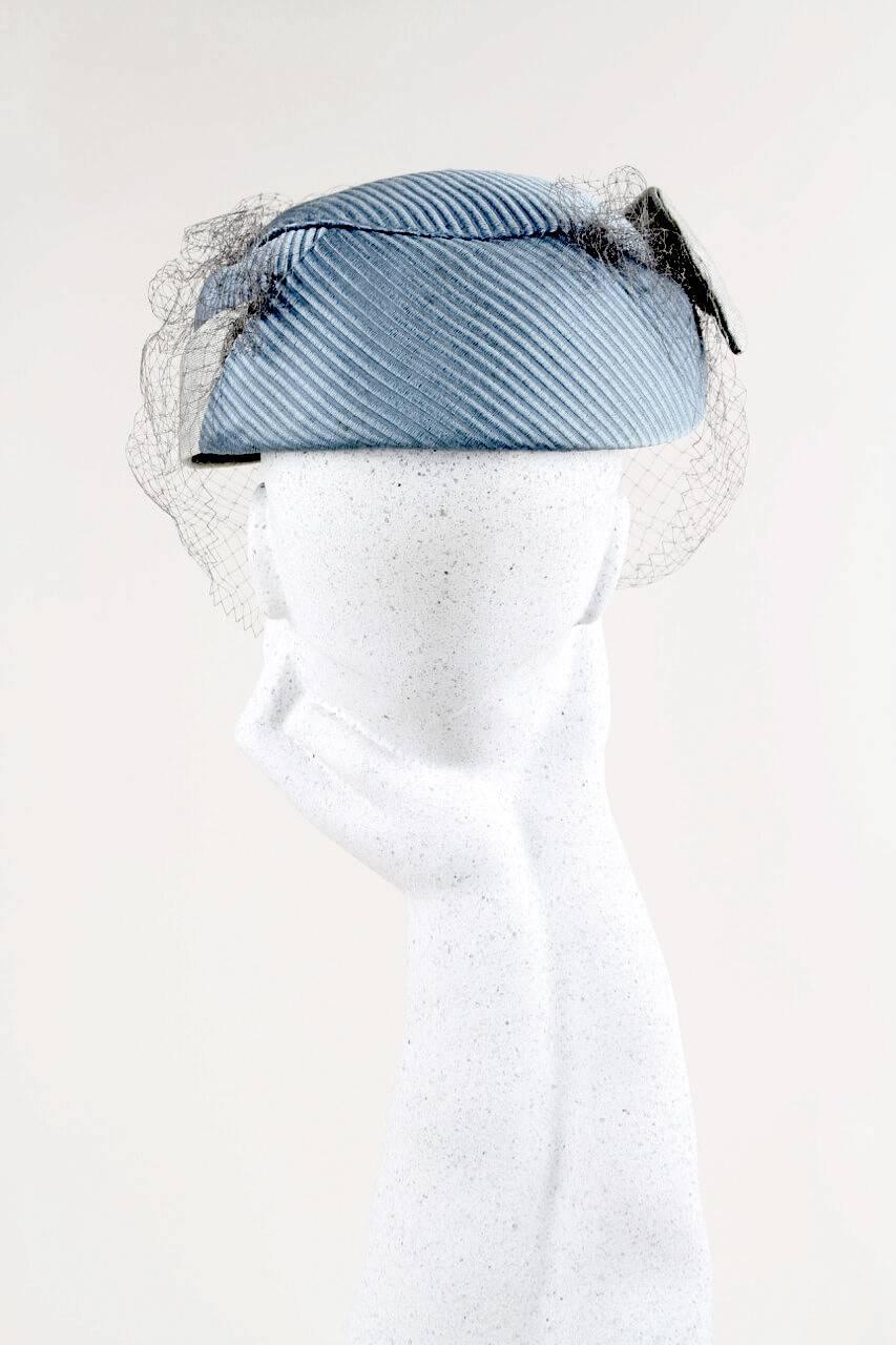 This ultra-elegant 1950s cocktail hat is designed from an aquamarine ribbed stiff fabric and a light grey veil. It is decorated with a silver fabric detailing that starts at the left side and comes out on the right side as a stiff little wing. It