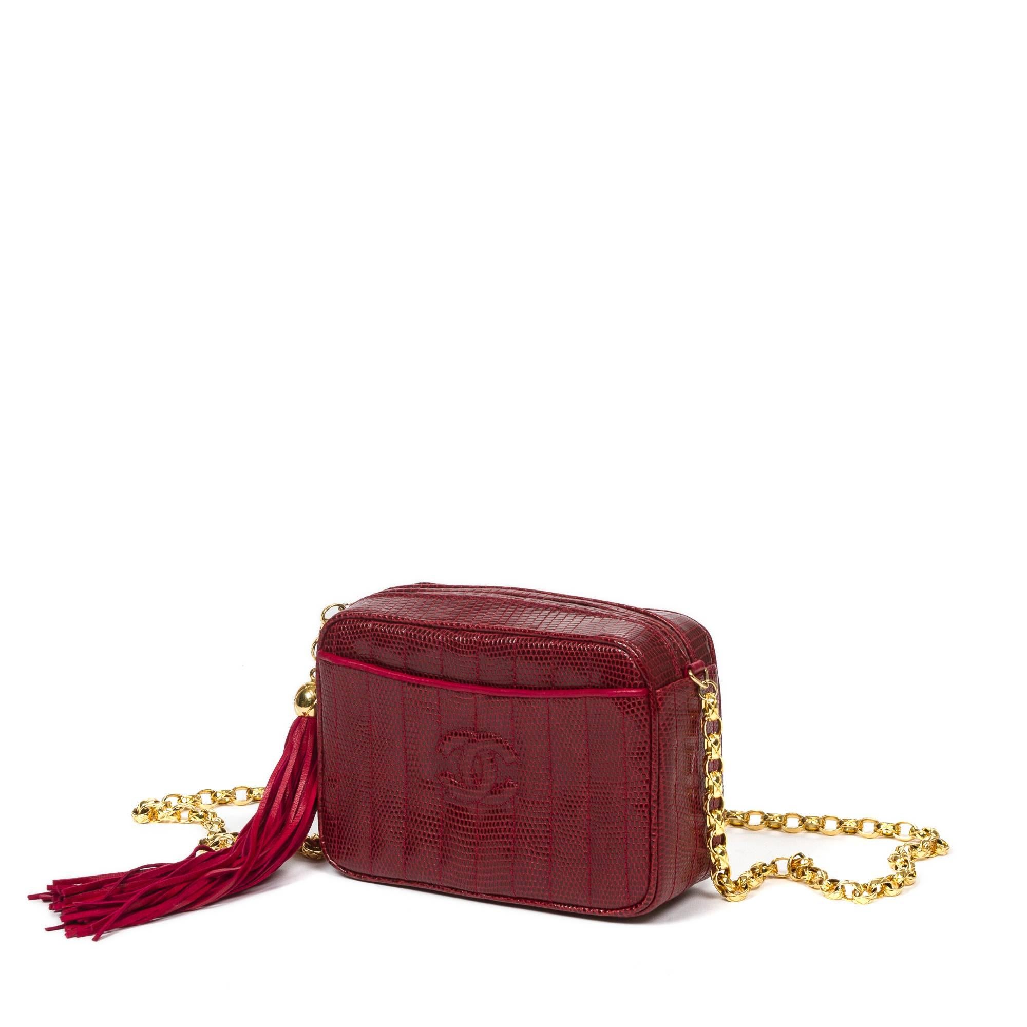 Vintage Tassel Camera Bag in garnet red lizard skin with thick link gold tone chain strap. 2 exterior slip pockets with red leather piping. Zip closure with red leather tassel zipper toggle. Black leather lined interior with one zip pocket. Gold