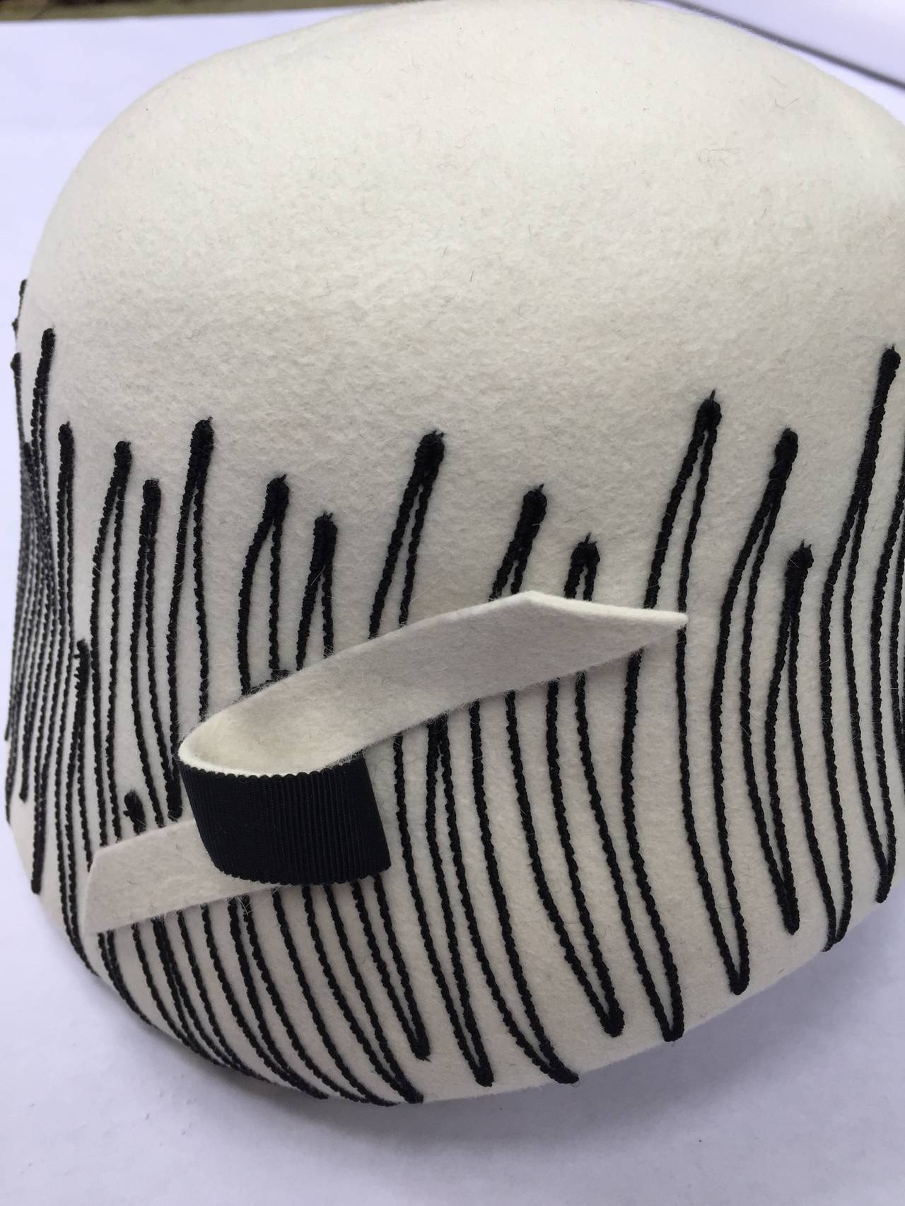 Elsa Schiaparelli 1950s off white Cloche-style wool felt hat.  The hat has a deep, cylindrical, domed crown of off-white or cream felt which extends downwards on one side. Around the crown is black zig-zag stitching, and on the side where the crown