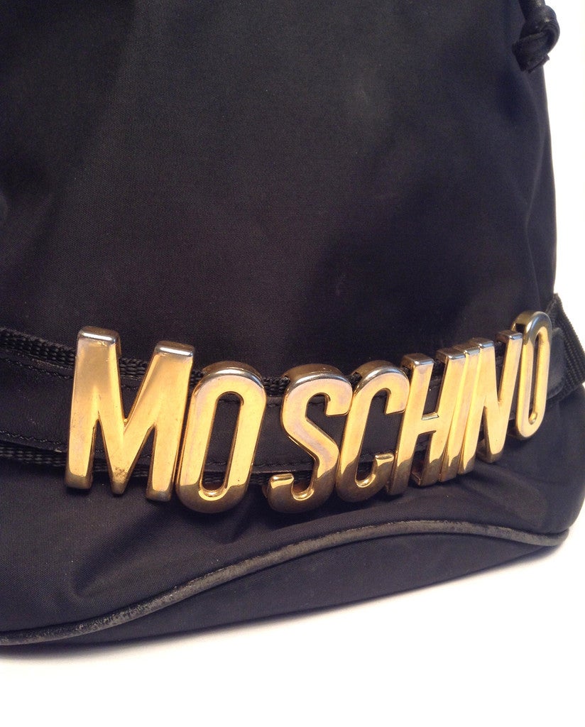 An amazing, every day carry-all with the classic Moschino gold-tone logo at front. The spacious and practical cross body features nylon body, and black leather trim with gold hardware. Drawstring closure. Adjustable shoulder strap with silver buckle