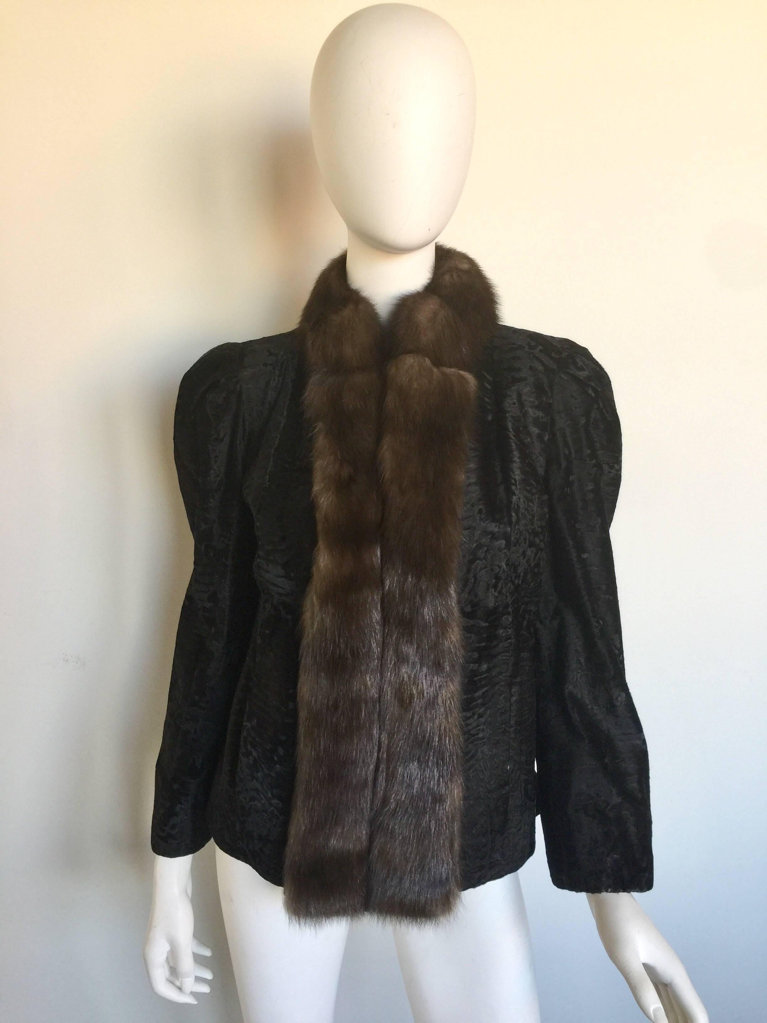 This jacket has a black broadtail lamb exterior embossed with a faint pattern. The front of the coat has a brown female mink fur lapel and collar. The coat is fully lined and has two front hidden pockets. The jacket has a hook enclosure and gathered