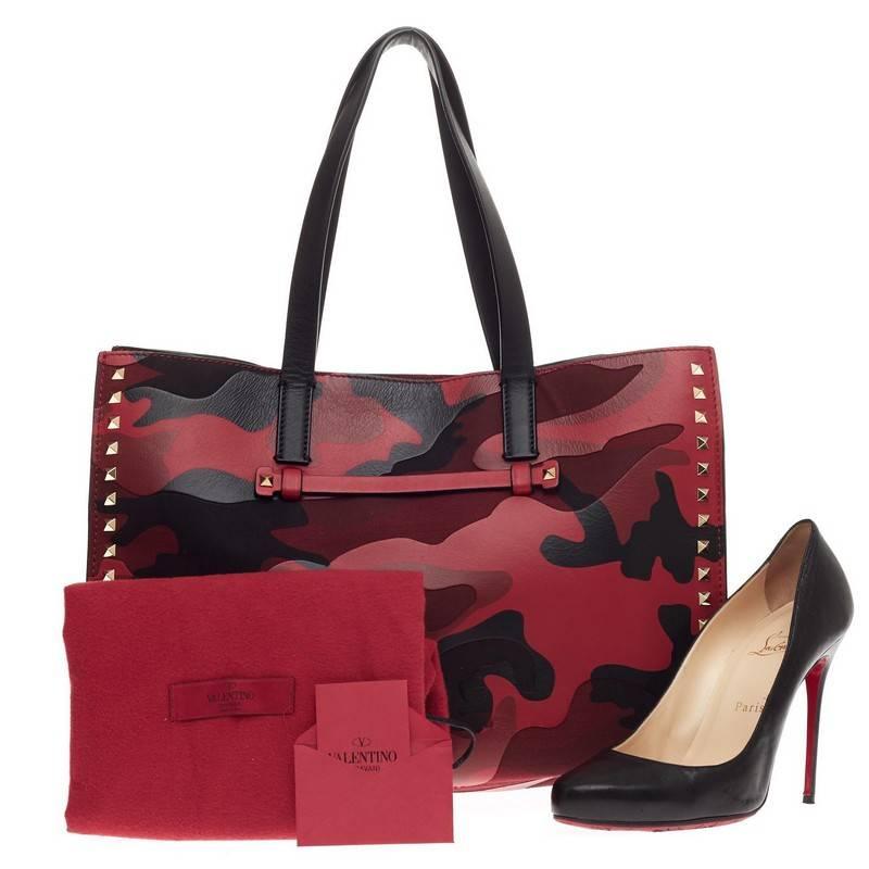 This authentic Valentino Rockstud Open Tote Camo Leather and Canvas Medium is a stylishly edgy bag that is one of today's most sought-after styles. Crafted from camouflage leather and canvas in shades of red, maroon and black, this eye-catching tote