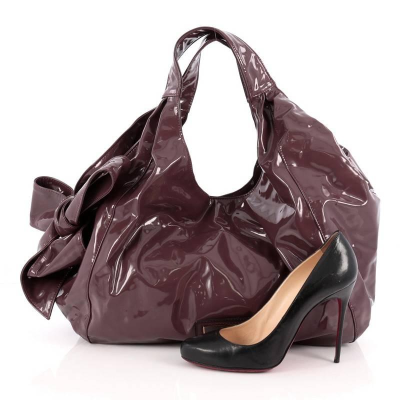 This authentic Valentino Nuage Bow Hobo Patent Large is a stylish, sleek hobo that showcases edgy femininity. Constructed from purple patent leather, this bag features large looping straps with oversized side bow detail and plated designer logo and