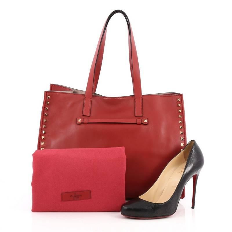This authentic Valentino Rockstud Open Tote Leather Medium is a stylishly edgy bag that is one of the most sought-after styles. Crafted from red leather, this eye-catching tote features dual flat tall leather handles, signature gold-tone pyramid