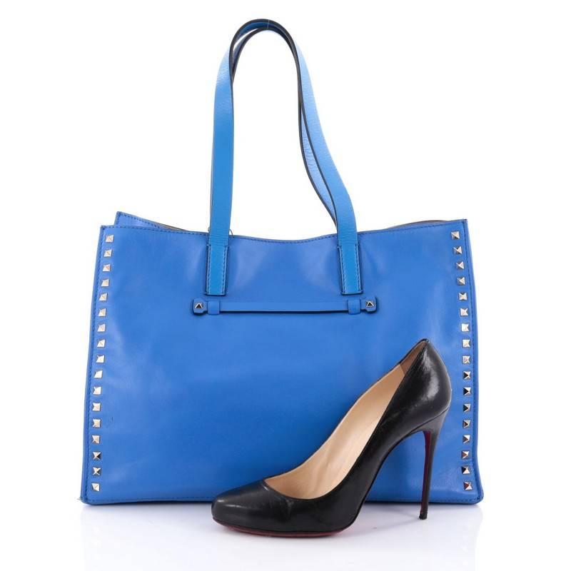 This authentic Valentino Rockstud Open Tote Leather Medium is a stylishly edgy bag that is one of the most sought-after styles. Crafted from blue leather, this eye-catching tote features dual flat tall leather handles, signature gold-tone pyramid