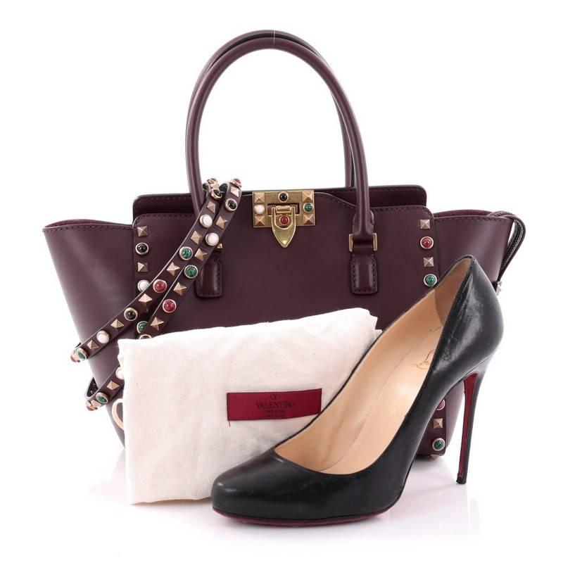 This authentic Valentino Rolling Rockstud Tote Leather with Cabochons Small mixes chic styling with luxurious flair. Crafted from burgundy smooth leather, this stand-out bag features dual-flat leather handles, colorful cabochon and semi-precious