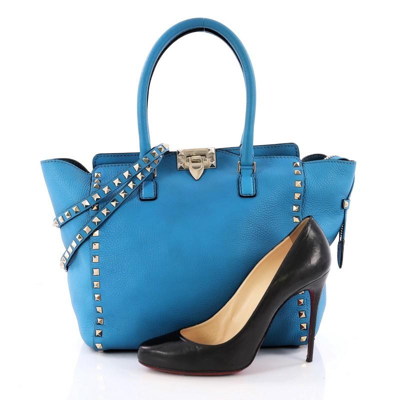This authentic Valentino Rockstud Tote Rigid Leather Medium is a stylish and iconic bag that is one of today's most sought-after styles. Crafted from beautiful blue rigid leather, this chic tote features signature gold pyramid stud borders,
