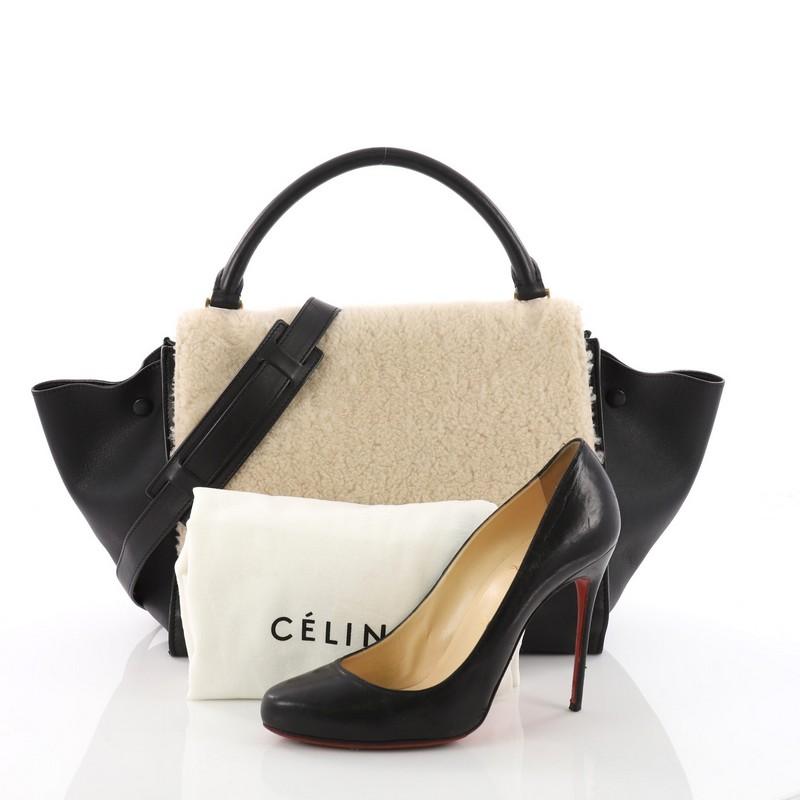 This authentic Celine Trapeze Handbag Shearling and Leather Medium is every fashionista's dream. Crafted in beige shearling and black leather, this classic bag features a top handle, exterior back pocket and gold-tone hardware accents. Its square