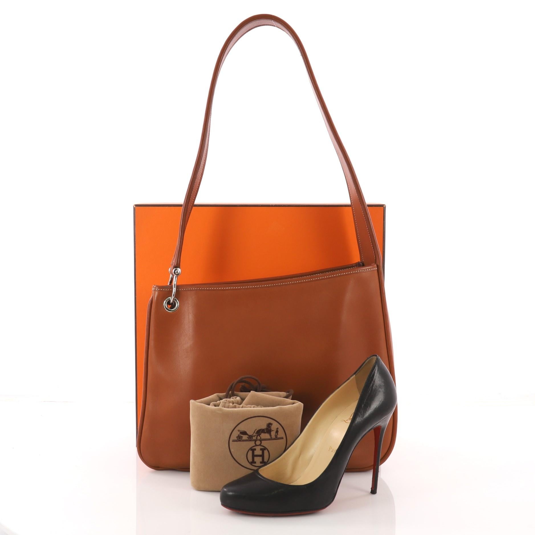 This Hermes Sac Manille II Handbag Leather, crafted in Fauve brown leather, features a wide looping shoulder strap and palladium-tone hardware. It opens to a brown leather interior with zip and slip pockets. **Note: Shoe photographed is used as a