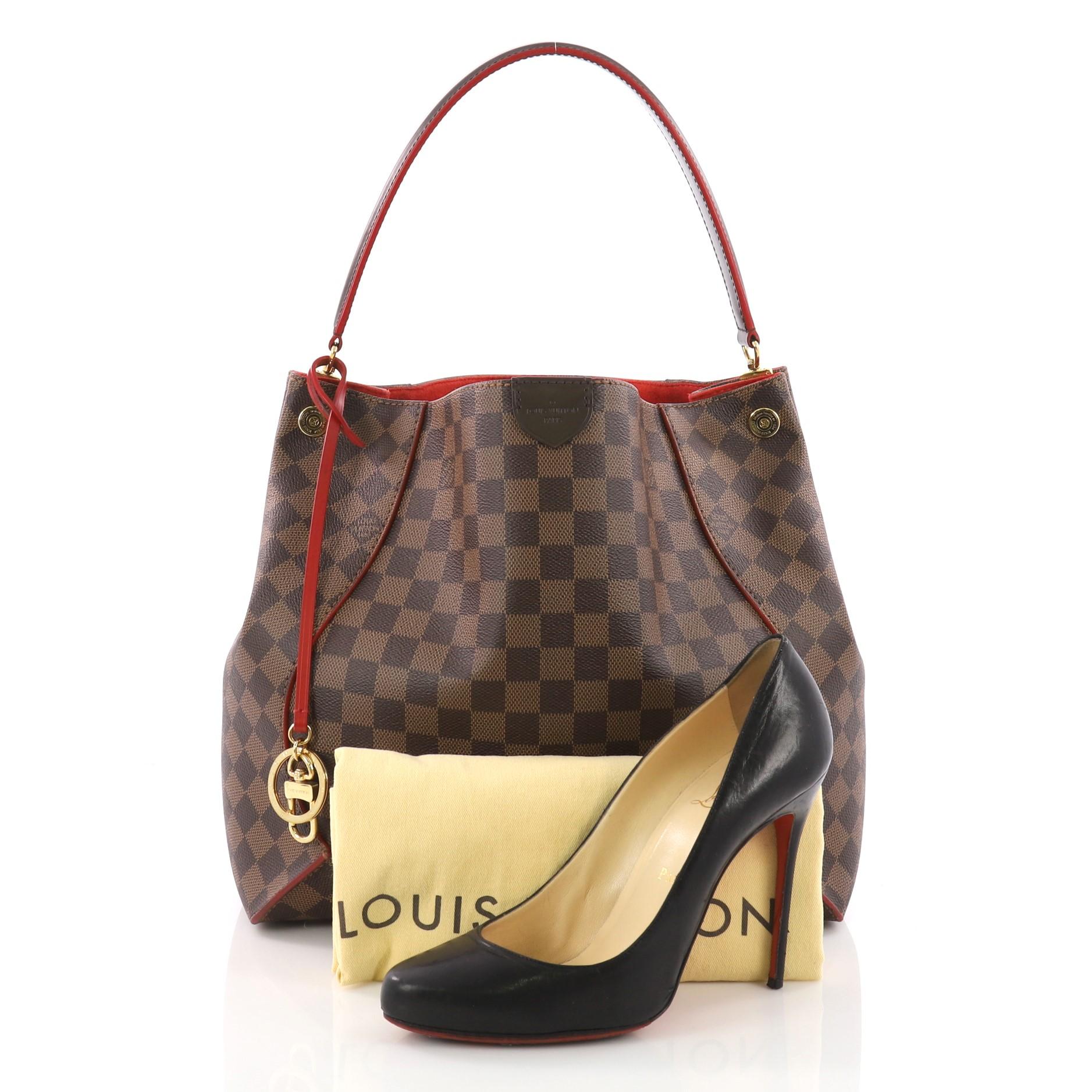 This Louis Vuitton Caissa Hobo Damier, crafted from damier ebene coated canvas, features a looping shoulder strap with polished gold anchors, cherry red leather trims, and gold-tone hardware accents. Its wide open top showcases a red microfiber