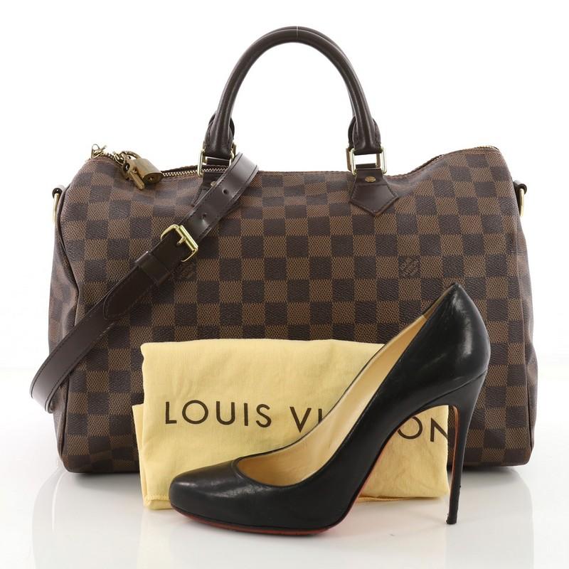 This Louis Vuitton Speedy Bandouliere Bag Damier 35, crafted in damier ebene coated canvas, features dual rolled handles, dark brown leather trims and gold-tone hardware. Its two way zip closure opens to a red fabric interior with slip pocket.