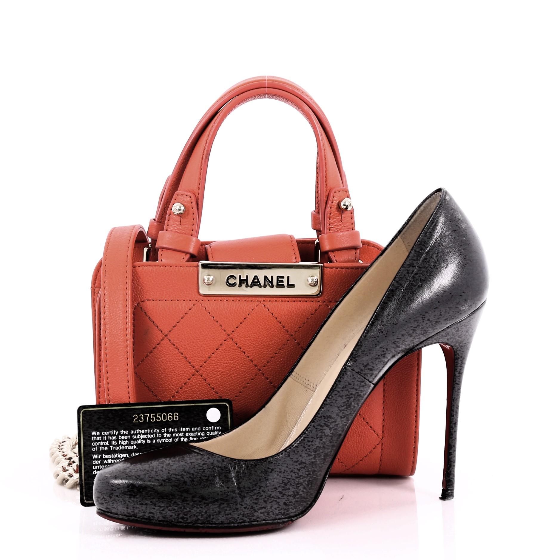 This Chanel Label Click Shopping Tote Quilted Calfskin Mini, crafted in red orange quilted calfskin leather, features dual flat leather handles, chunky chain link strap with leather pads, and gold-tone hardware. Its magnetic snap closure opens to a