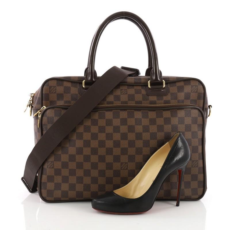 This Louis Vuitton Icare Laptop Bag Damier, crafted from damier ebene coated canvas, features a structured, boxy silhouette, dual rolled leather handles, front zip pocket and gold-tone hardware. Its zip closure opens to a brown fabric interior.