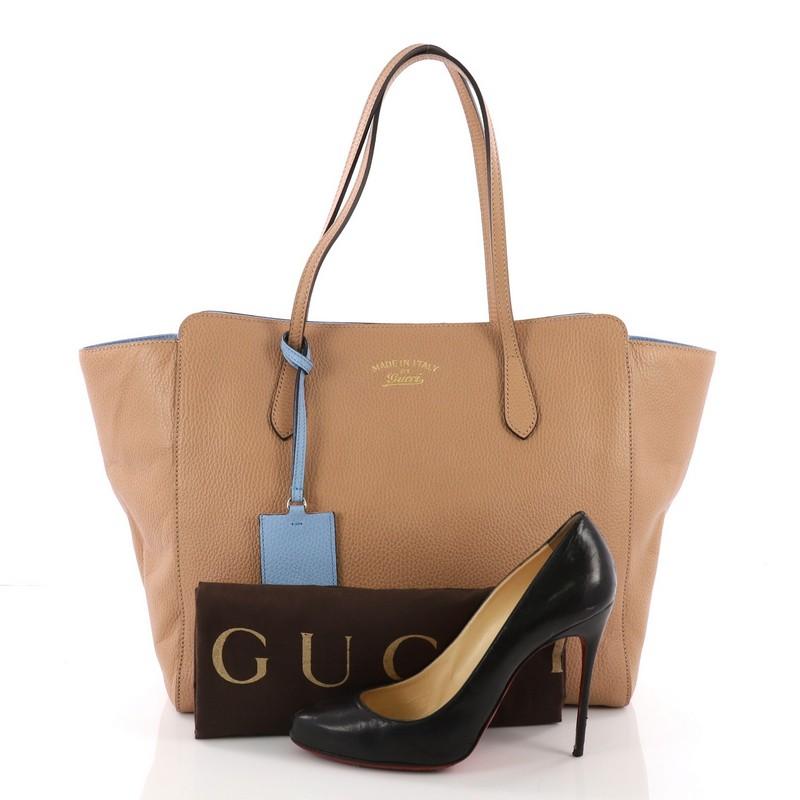 This Gucci Swing Tote Leather Medium, crafted in nude leather, features tall dual-slim handles, Gucci stamped logo at the front, and silver-tone hardware. Its hidden magnetic snap closure opens to a beige fabric interior and includes side zip and