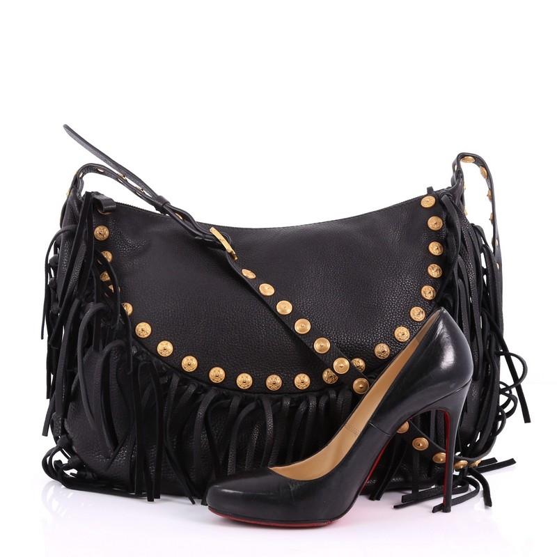 This Valentino C-Rockee Fringe Hobo Studded Leather Large, crafted from black leather, features hand-knotted long leather fringes, gryphon embossed studs, adjustable flat leather handle with stud detailing, whipstitched base, and gold-tone hardware.