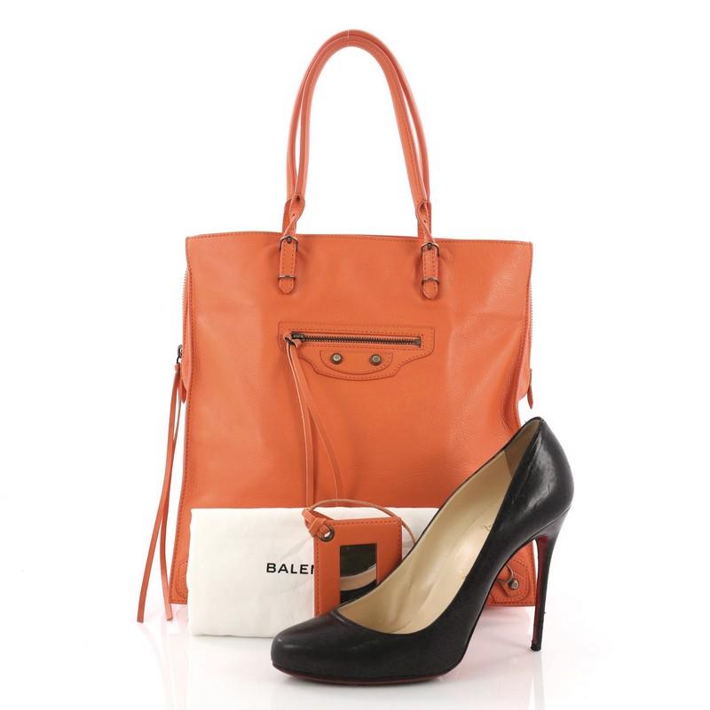This Balenciaga Papier Classic Studs Zip Tote Leather Medium, crafted from orange leather, features dual rolled handles with buckle details, Balenciaga's iconic classic studs, exterior front zip pocket, and aged brass-tone hardware. It opens to an