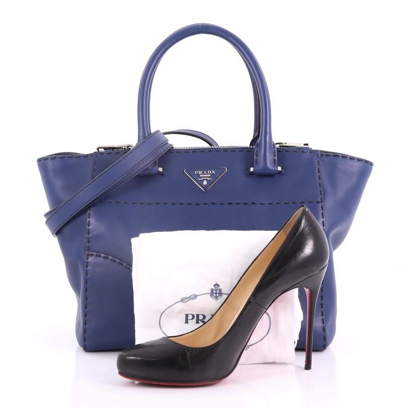 This Prada Twin Pocket Tote Stitched City Calfskin Small, crafted from blue calfskin leather, features dual-rolled handles, expanded winged sides, raised Prada logo at the center, and silver-tone hardware. Its magnetic snap closure opens to a beige
