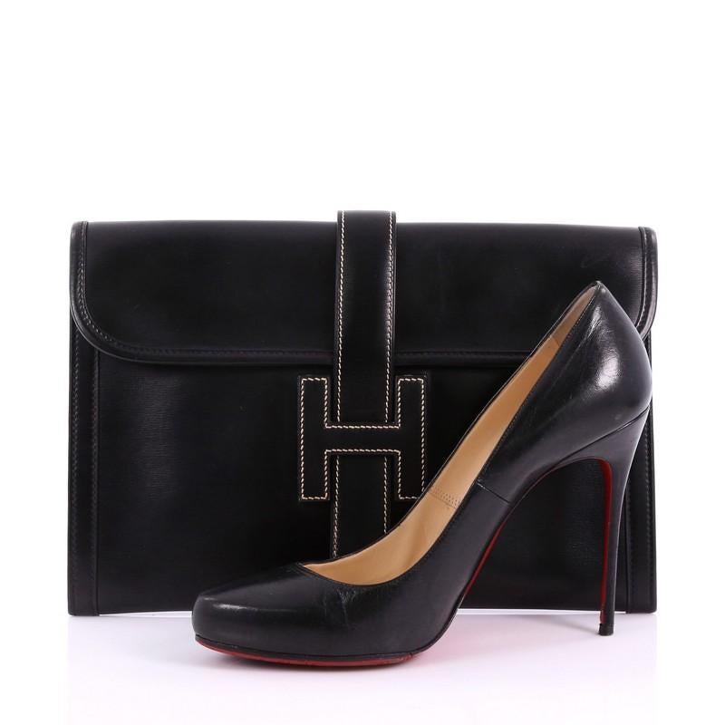 This Hermes Jige Clutch Box Calf PM, crafted in black noir box calf leather, features a cross-over flap and strap that tucks under the logo H. It opens to a beige fabric interior. Date stamp reads: I Circle (1979). **Note: Shoe photographed is used