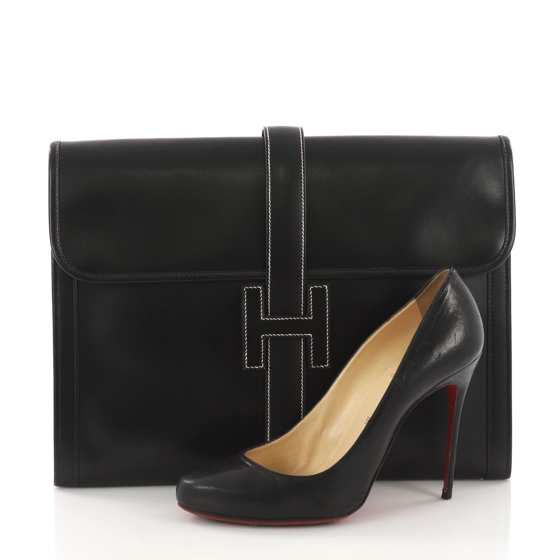 This Hermes Jige Clutch Box Calf GM, crafted in black calf leather, features a cross-over flap and strap that tucks under the logo H. It opens to a beige fabric interior. **Note: Shoe photographed is used as a sizing reference, and does not come